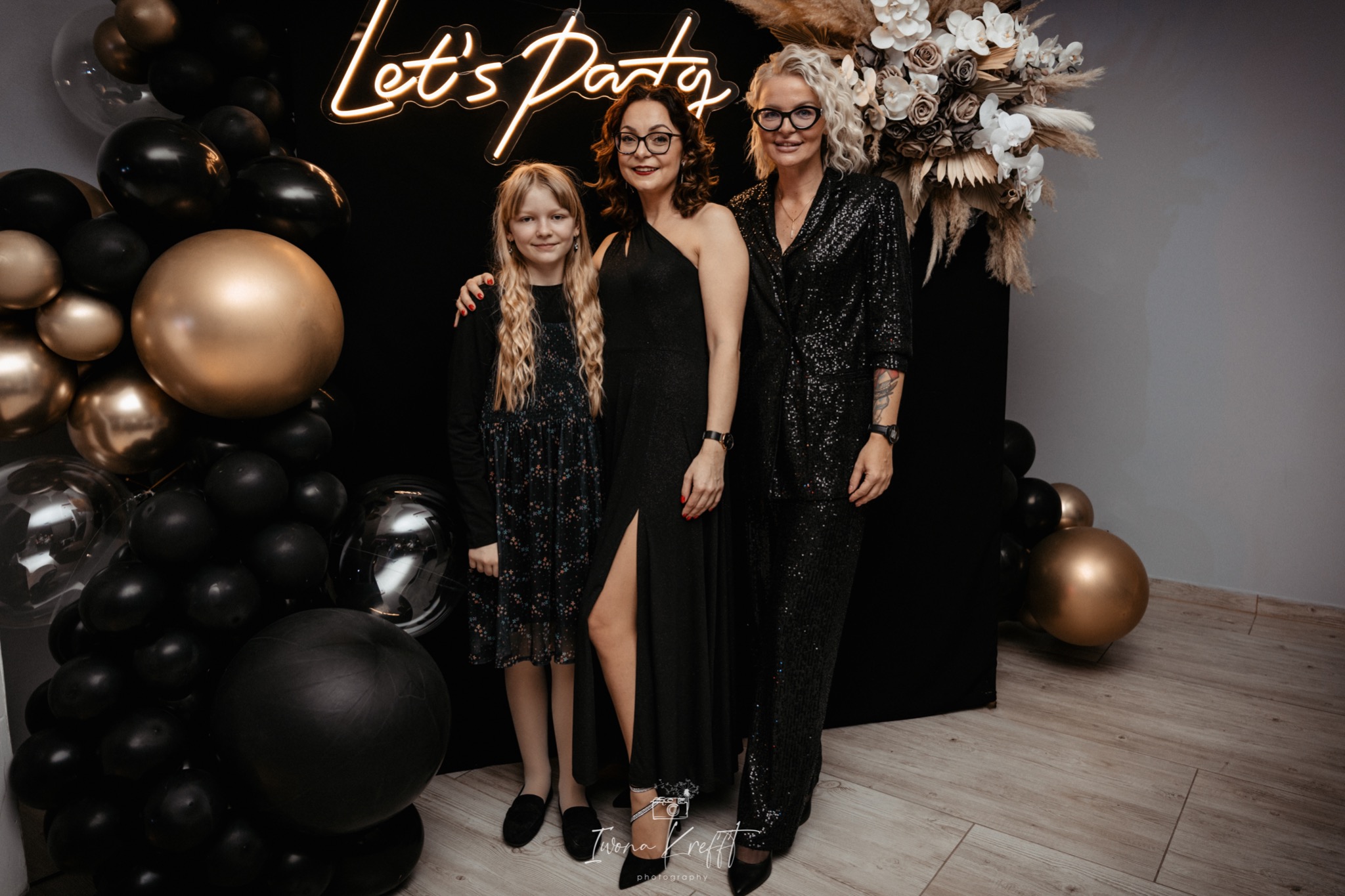 Trzy kobiety w eleganckich, czarnych strojach pozują na tle dekoracji z czarnych i złotych balonów oraz neonu 'Let's Party' w Sulęczynie. Kompozycja kwiatowa z pampasów dodaje uroku.