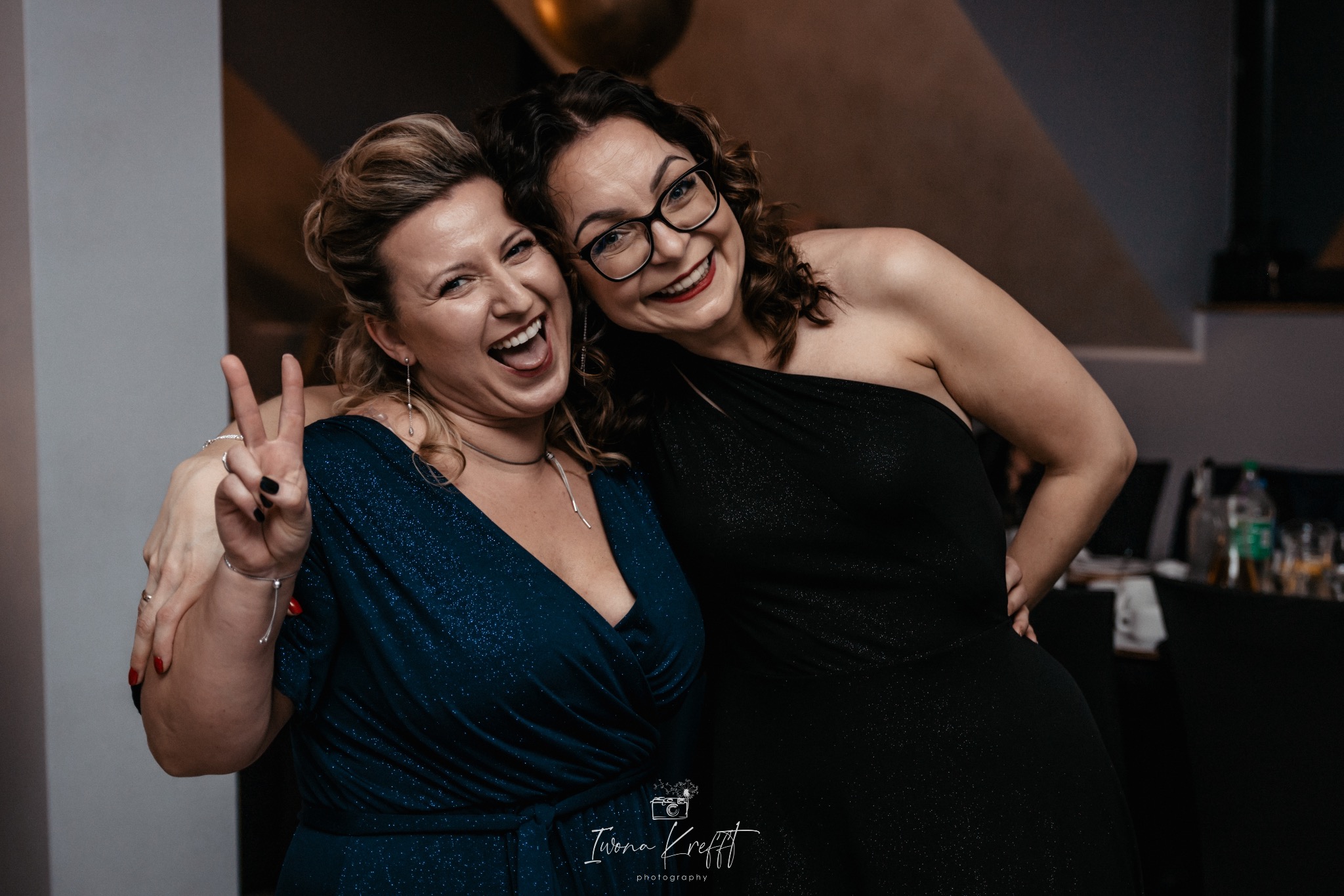 Dwie roześmiane kobiety w eleganckich sukienkach, jedna z uniesioną dłonią pokazująca znak zwycięstwa, zdjęcie zrobione w pomieszczeniu z ciemnym tłem.