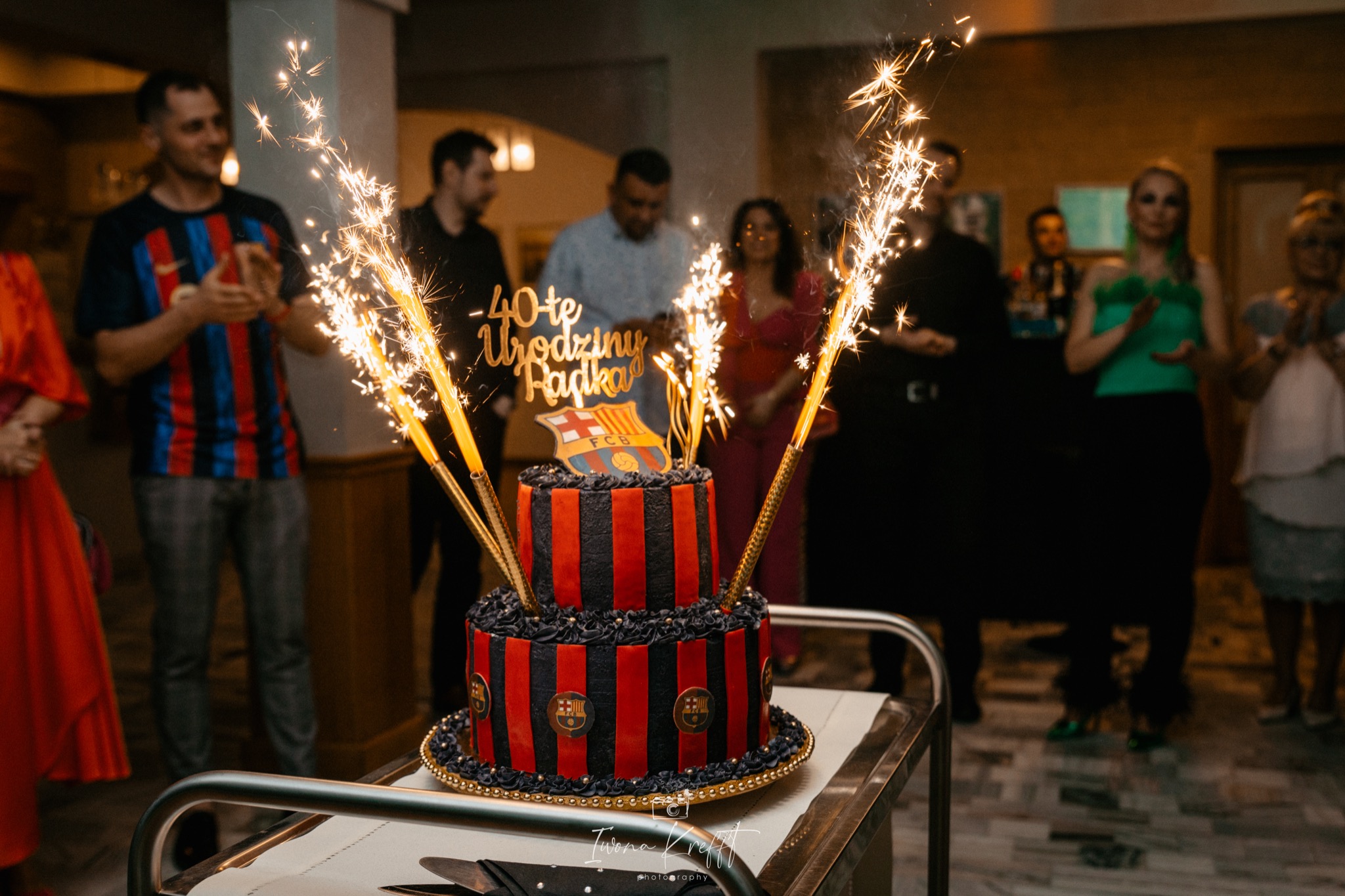 Dwupoziomowy tort urodzinowy w barwach FC Barcelony, z dekoracją '40-te Urodziny Radka' i fontannami iskier, na tle rozmytych sylwetek gości.