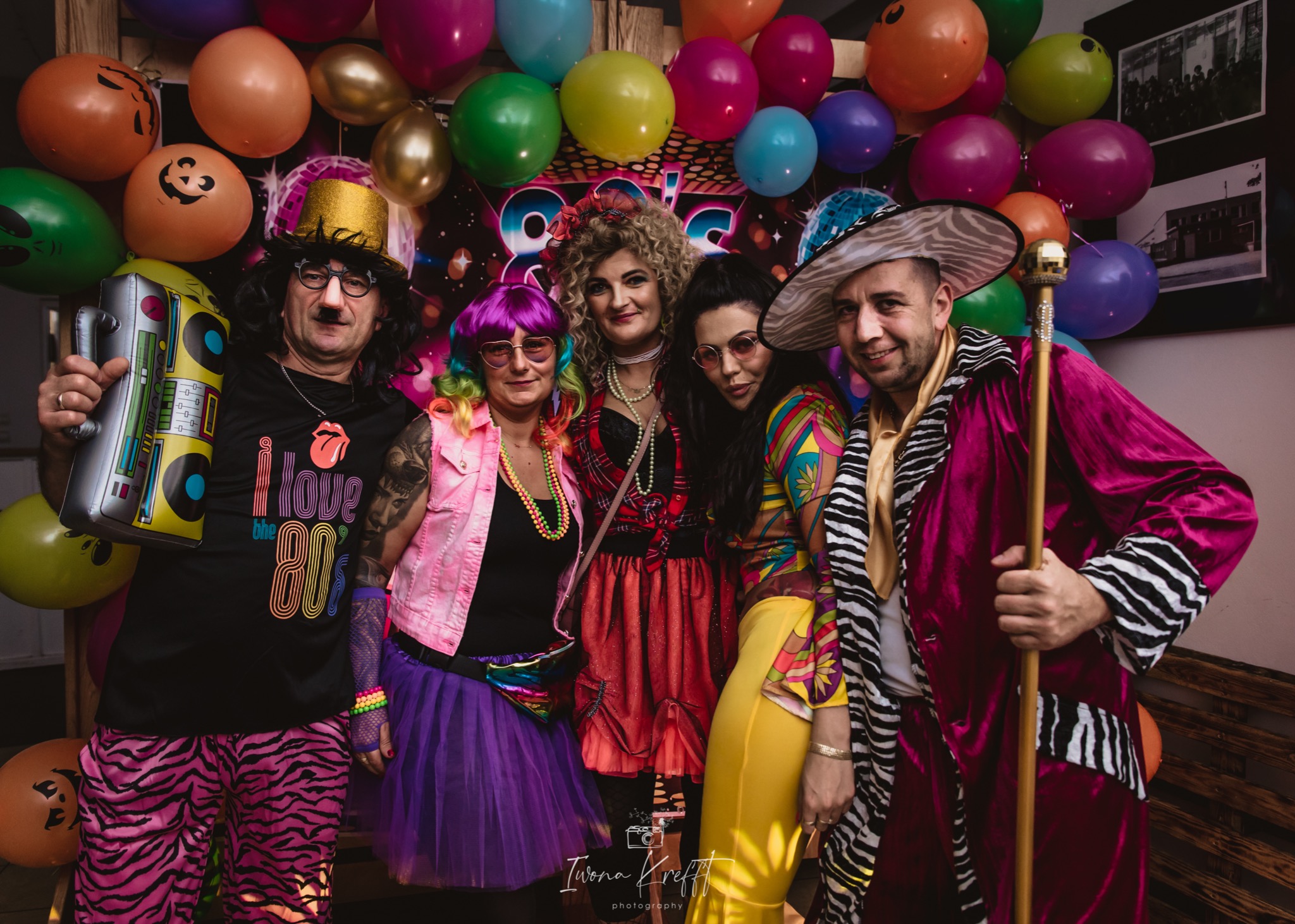Grupa osób przebranych w kolorowe stroje w stylu lat 80. pozuje na tle balonów podczas imprezy tematycznej, mężczyzna z wąsami trzyma nadmuchiwany magnetofon.