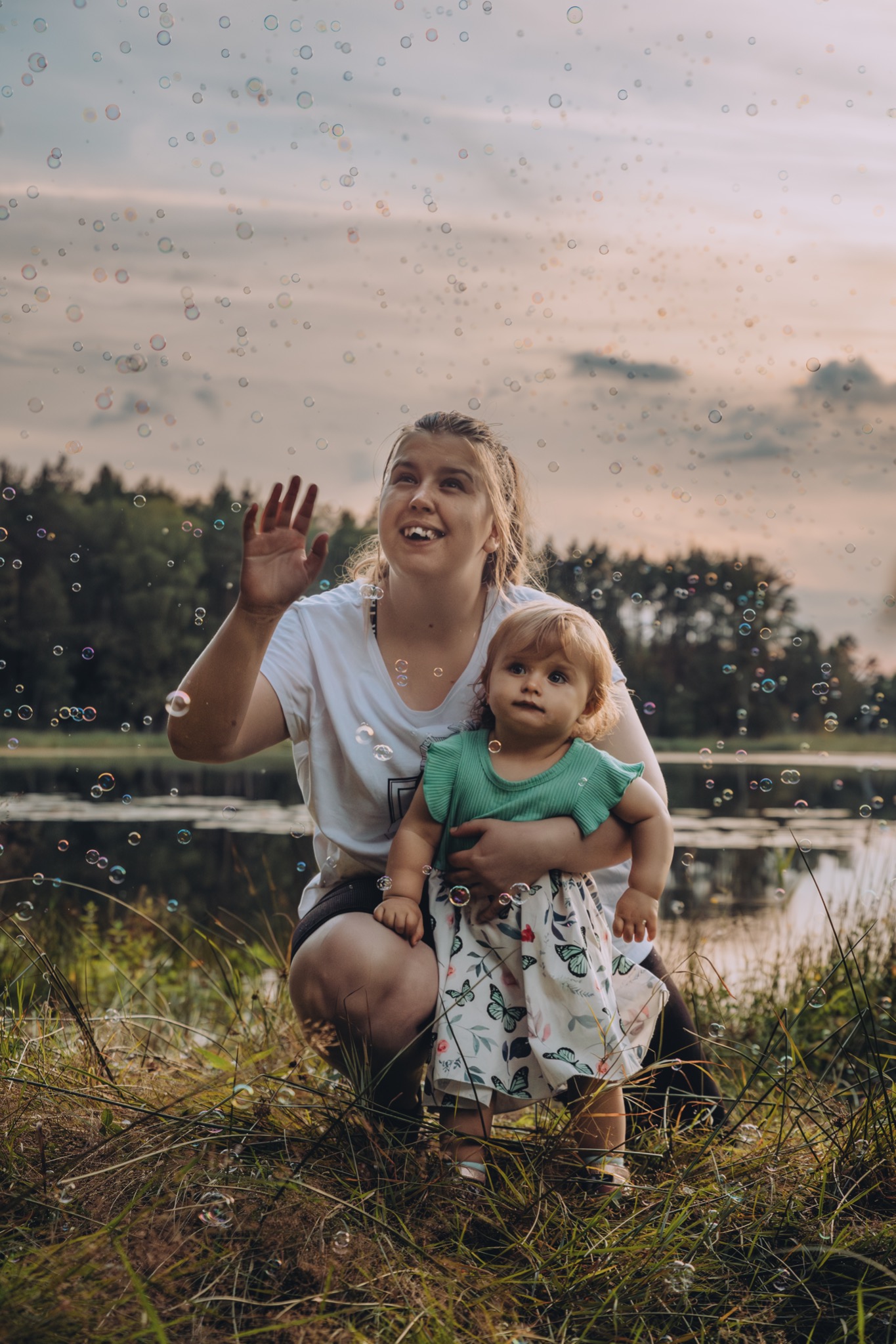 Kobieta kuca obok jeziora, trzymając małą dziewczynkę, w otoczeniu unoszących się baniek mydlanych, z uśmiechem wyciąga rękę w ich stronę.