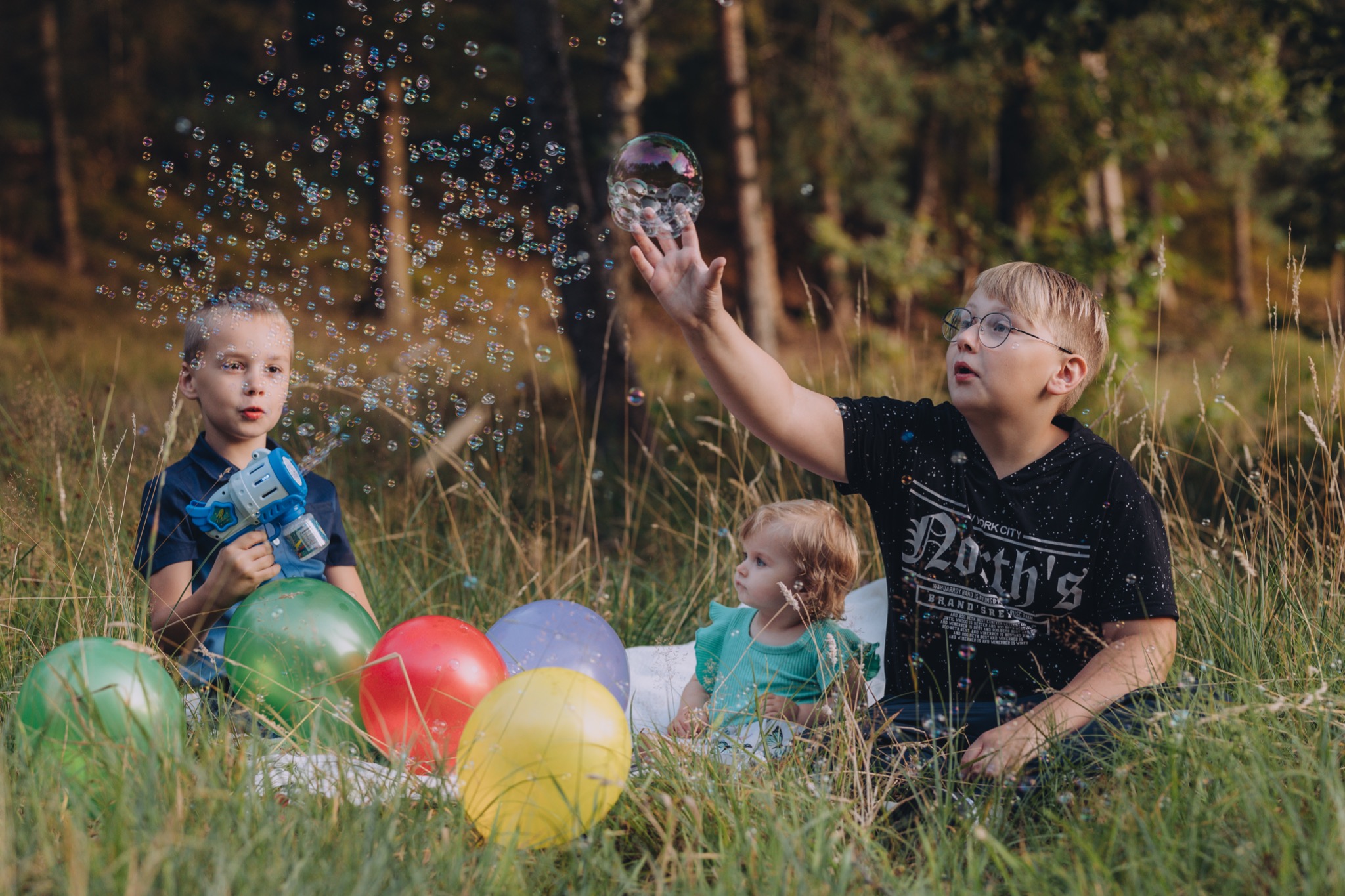 Trójka dzieci bawi się na łące z bańkami mydlanymi i kolorowymi balonami; chłopiec w okularach próbuje złapać bańkę, drugi chłopiec trzyma pistolet do robienia baniek, a mała dziewczynka siedzi obok.