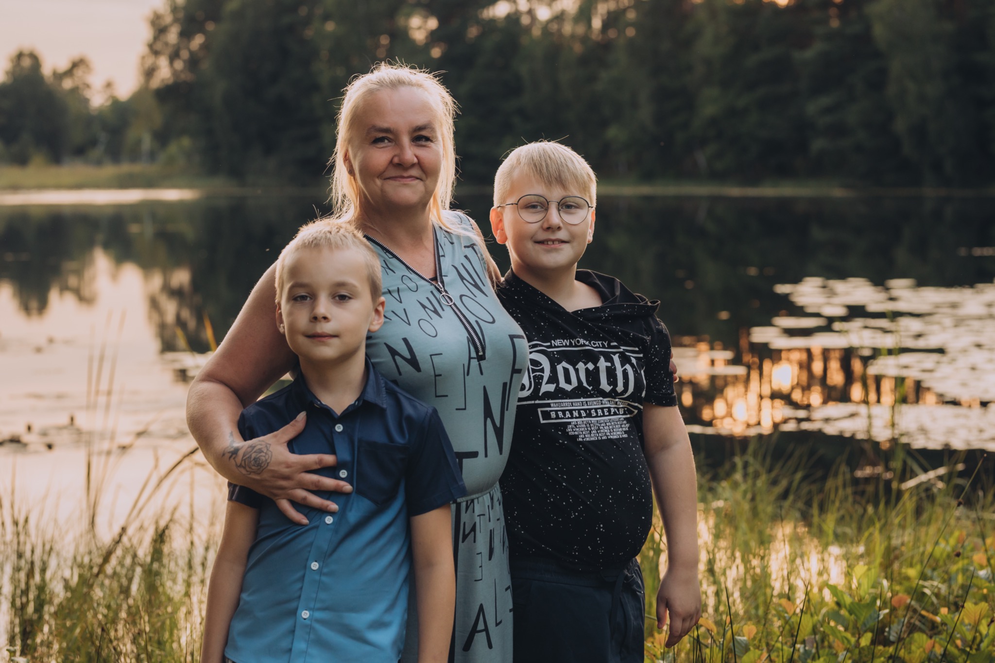 Portret kobiety z dwoma synami na tle jeziora i lasu o zmierzchu, widoczny tatuaż róży na dłoni kobiety.