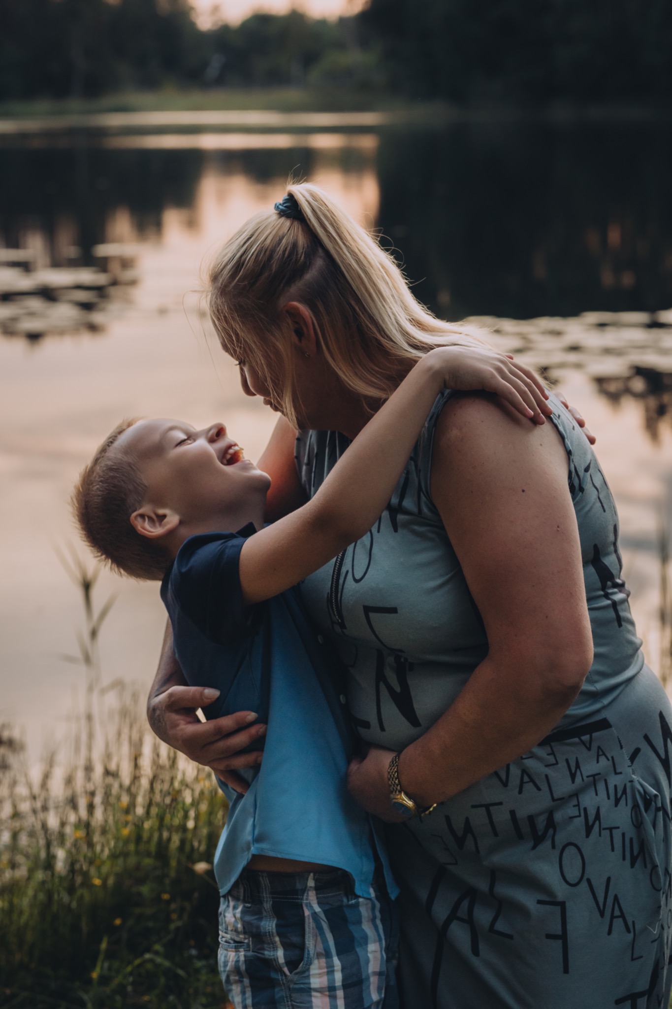 Uśmiechnięty chłopiec w niebieskiej koszuli mocno obejmuje blondwłosą kobietę w szarej sukience na tle jeziora i zieleni.