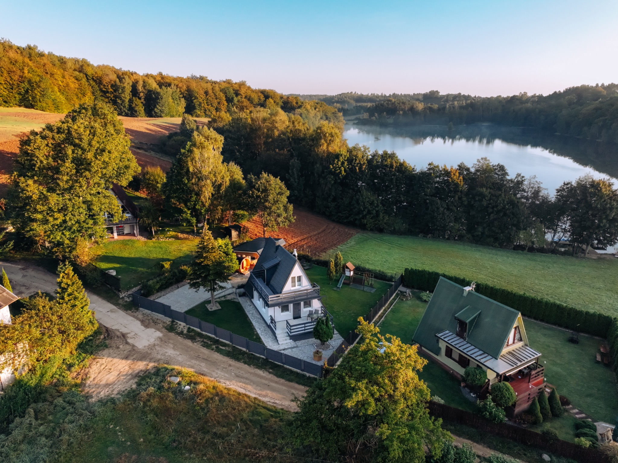 Zdjęcie z drona przedstawia malowniczy krajobraz z domami jednorodzinnymi, jeziorem w tle oraz otaczającą je bujną zielenią drzew i pól oświetlonych porannym słońcem.