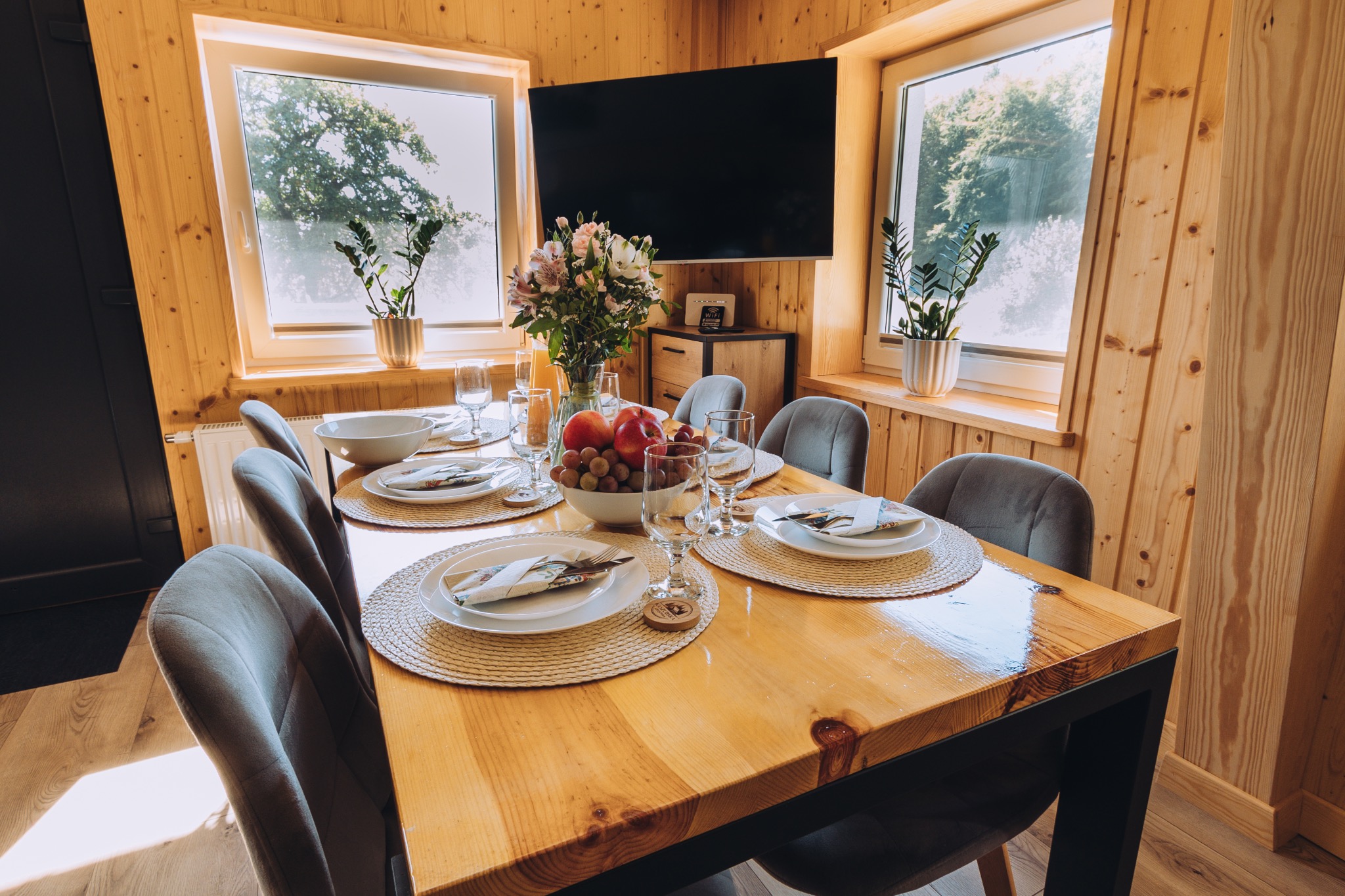 Stół z drewna z nakryciem dla sześciu osób, kwiaty w wazonie i owoce w misie, drewniane ściany i okna z widokiem na zewnątrz, telewizor na ścianie.