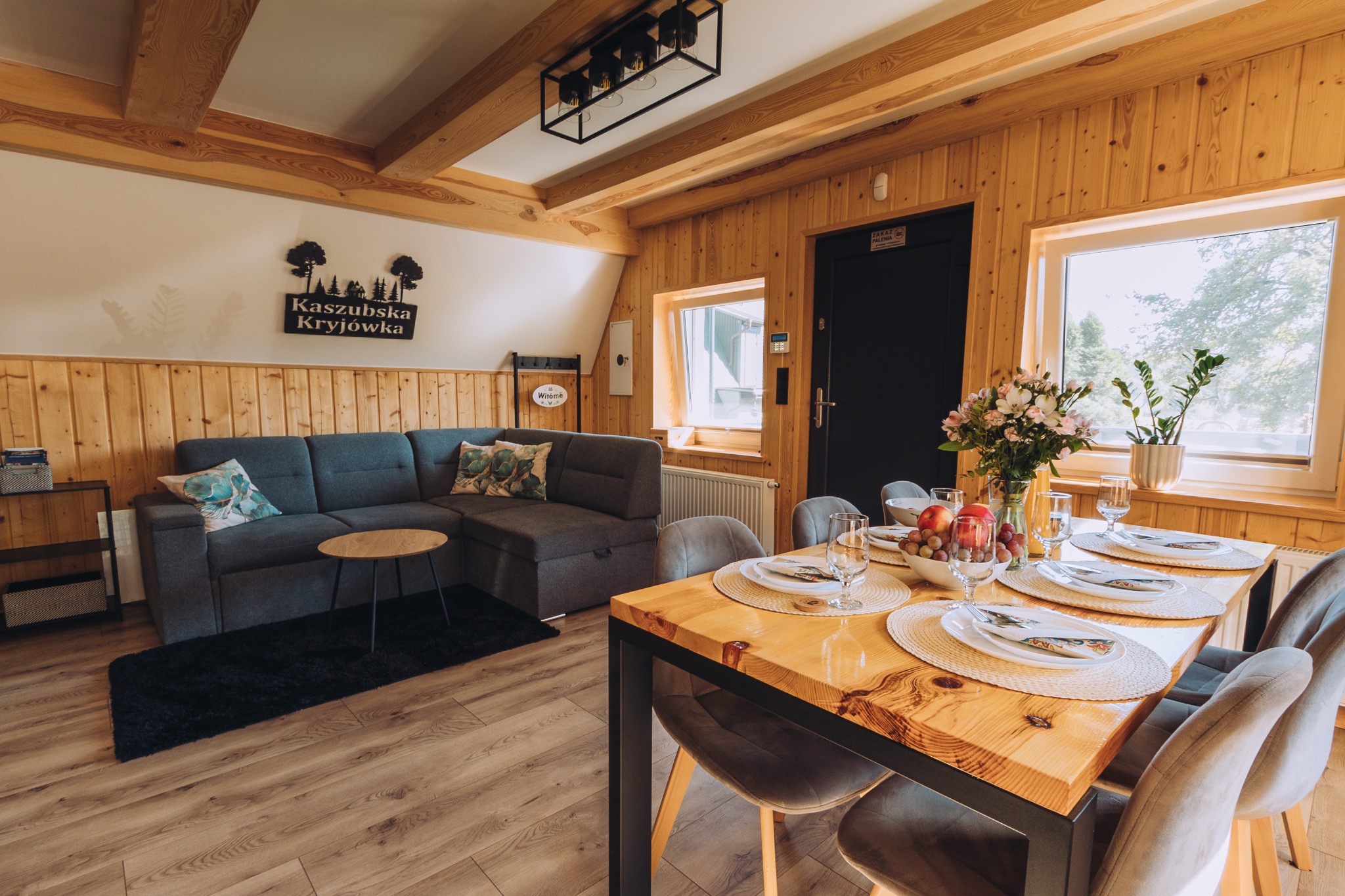 Jasne wnętrze w stylu rustykalnym z drewnianymi panelami, szarym narożnikiem, stołem z nakryciem i dekoracjami, widok na okno z roślinami i tabliczką z nazwą miejsca.