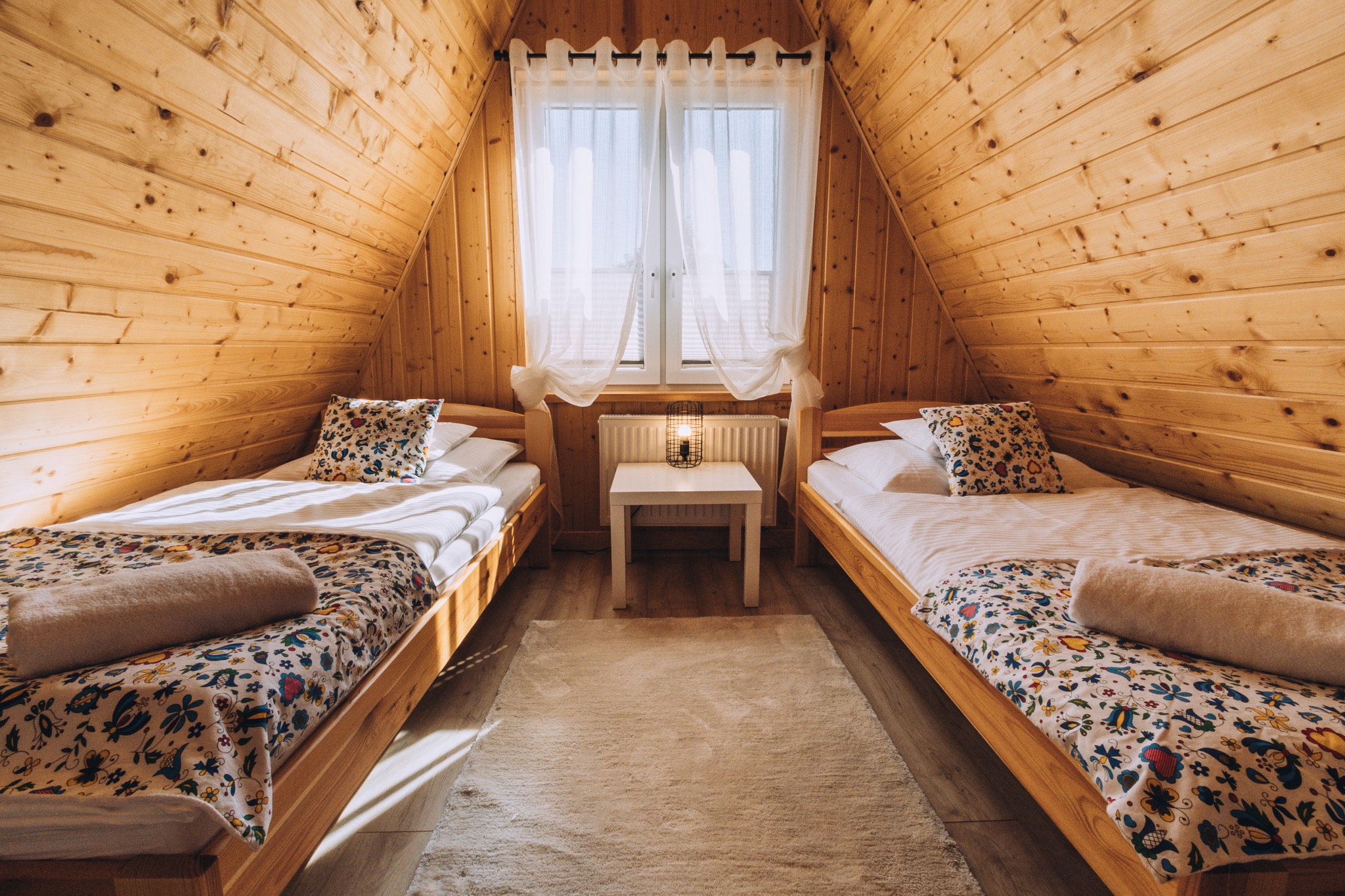 Drewniany pokój na poddaszu z dwoma pojedynczymi łóżkami, wzorzystą pościelą i dywanem, oświetlony lampką na stoliku nocnym pod oknem.