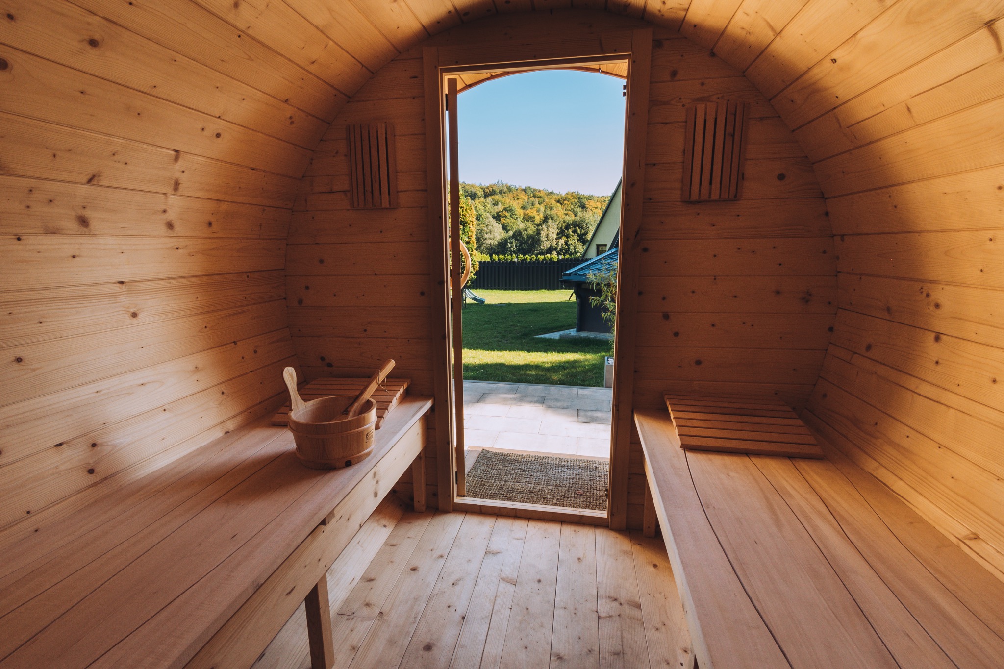 Wnętrze drewnianej sauny beczki z widokiem na ogród przez otwarte drzwi, drewniane ławki po bokach, wiadro z czerpakiem.