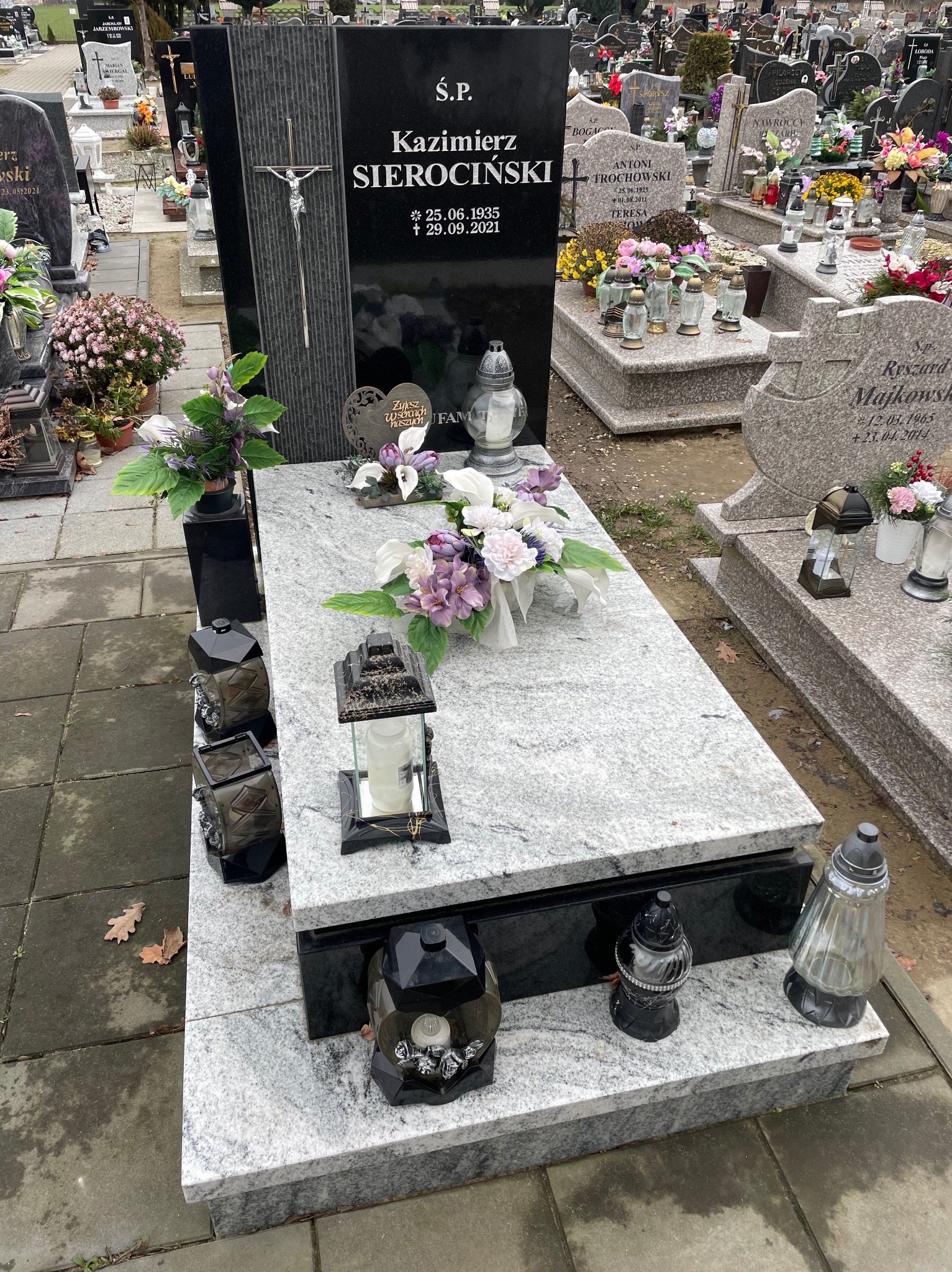 Granitowy nagrobek z czarną tablicą, krzyżem i inskrypcją, ozdobiony kwiatami i lampionami, na tle cmentarza z innymi nagrobkami.