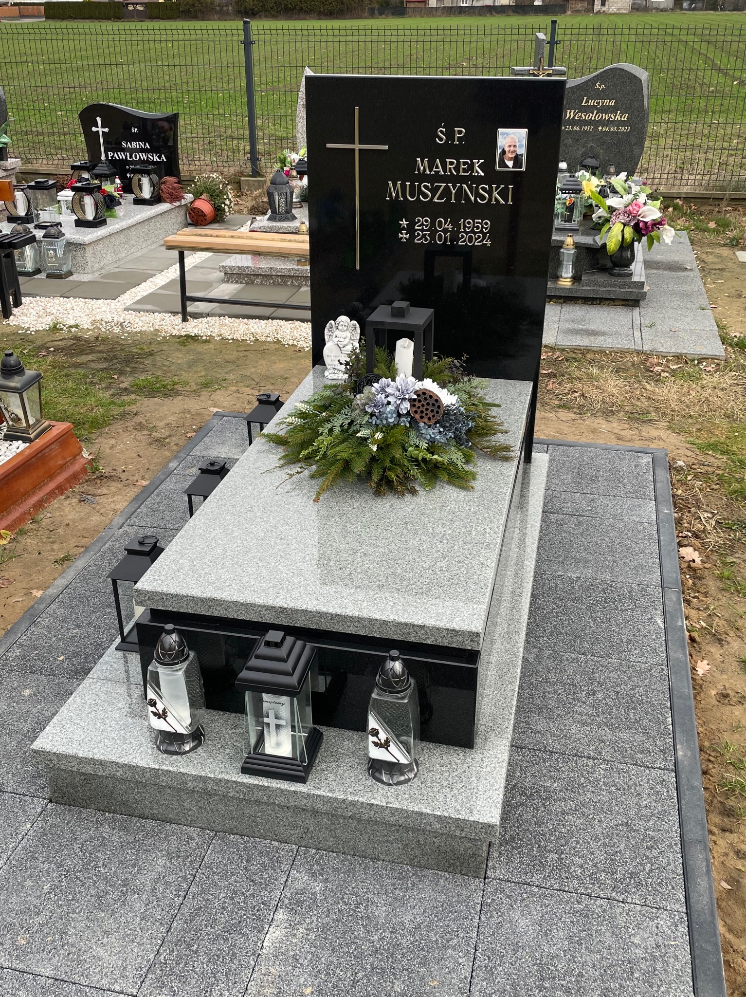 Granitowy nagrobek z czarną tablicą, ozdobiony wieńcem z kwiatów i lampionami na cmentarzu.