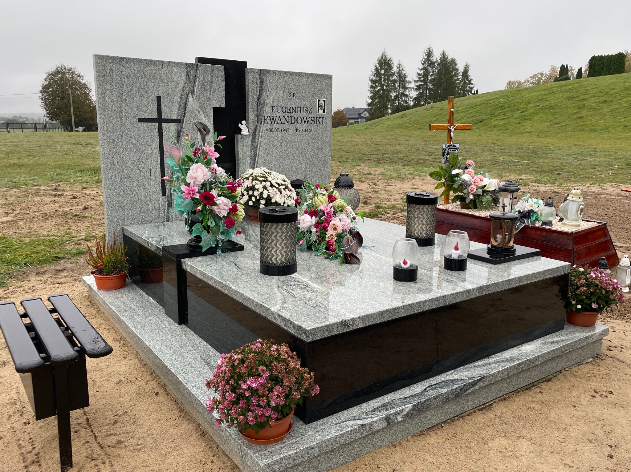 Granitowy nagrobek z czarnymi elementami, kwiatami i lampionami na cmentarzu w pochmurny dzień, z widocznym zdjęciem zmarłego Eugeniusza Lewandowskiego.