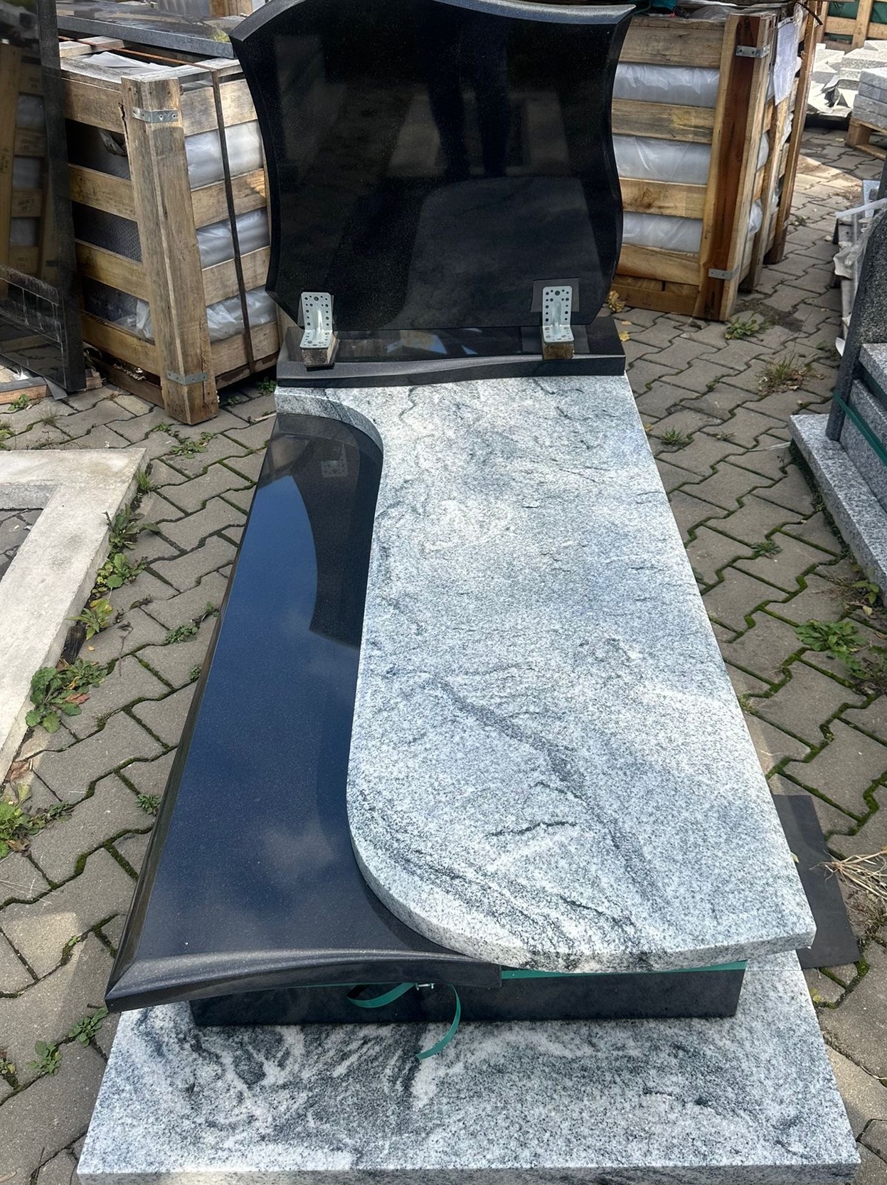 Nowoczesny nagrobek z czarnego i szarego granitu o falistym kształcie, z metalowymi uchwytami na znicze, na tle kostki brukowej i palet.