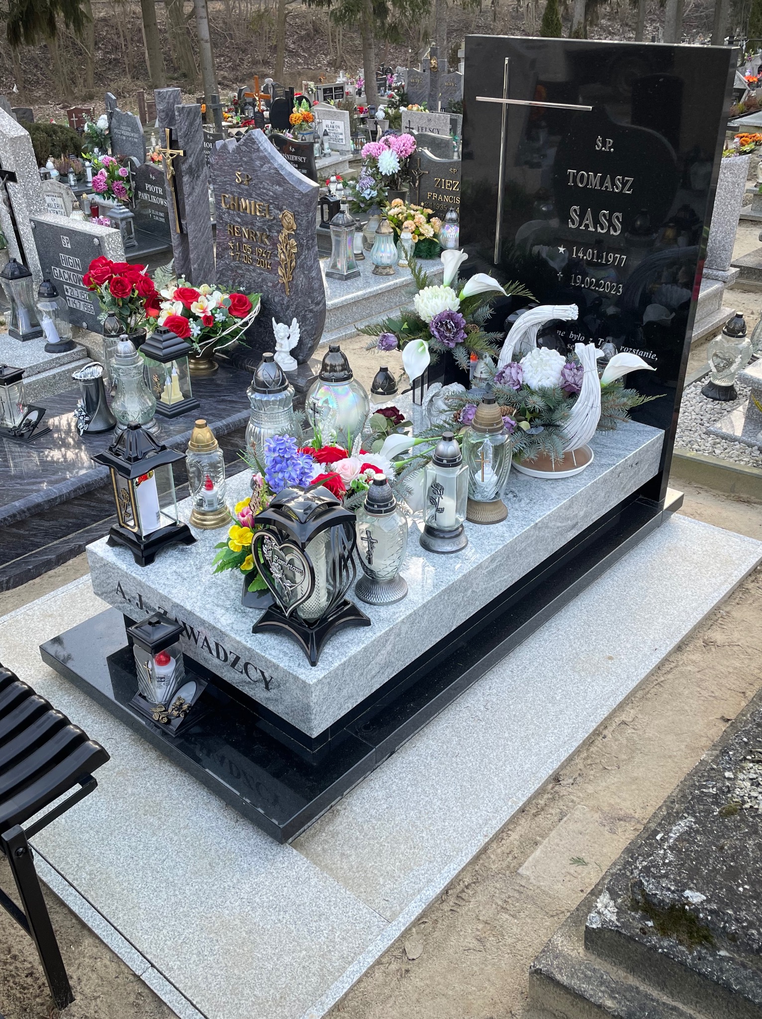 Czarny nagrobek z krzyżem i napisem Tomasz Sass, otoczony kwiatami i zniczami na cmentarzu, z widocznym imieniem A.J. Zwadczy na podstawie.