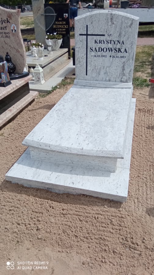 Jasny, granitowy nagrobek z krzyżem i inskrypcją 'Krystyna Sadowska', umieszczony na piaskowym podłożu. Widoczne inne nagrobki w tle.