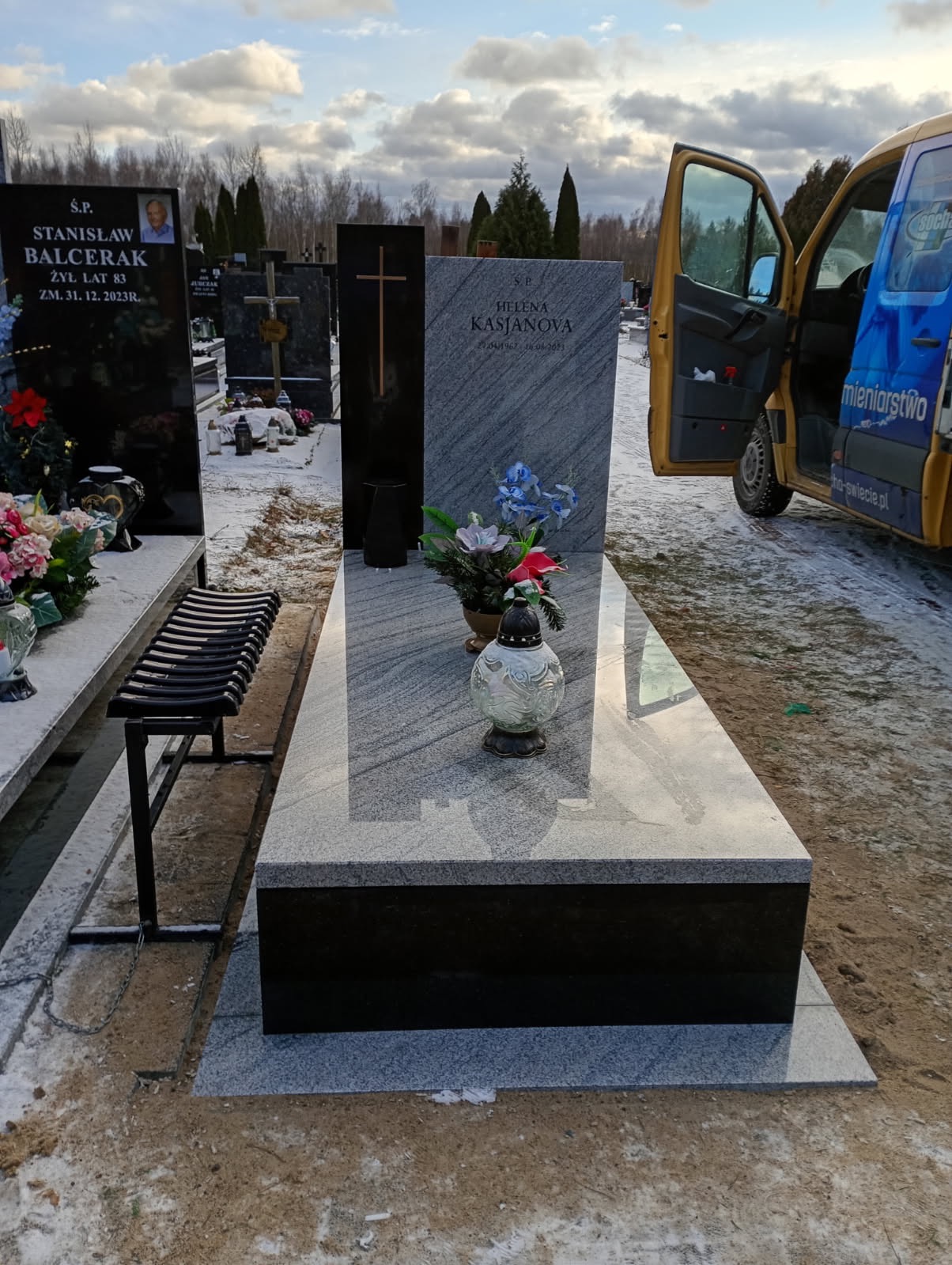 Jasnoszary nagrobek z ciemną podstawą i kwiatami na cmentarzu, z otwartymi drzwiami żółtego samochodu w tle. Widok na nagrobki i drzewa pod pochmurnym niebem.