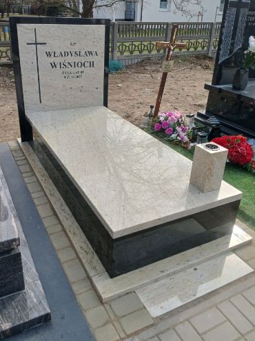 Jasny, dwukolorowy nagrobek z krzyżem i inskrypcją 'Ś.P. Władysława Wiśnioch' na płycie. Widoczne kwiaty i znicz. Tło stanowi ogrodzenie i zieleń.