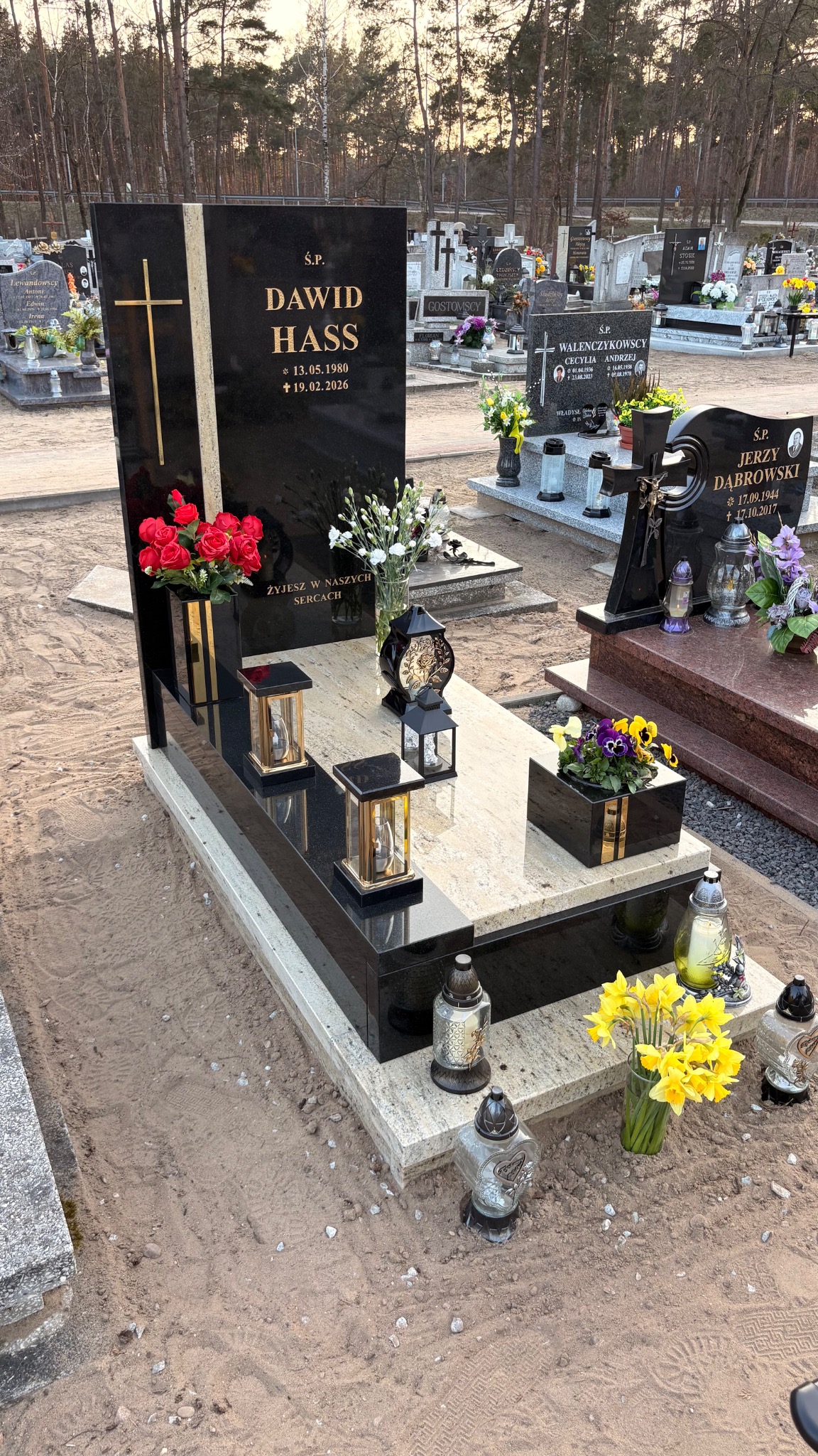 Czarny nagrobek z jasnym blatem, zdobiony złotym krzyżem i kwiatami. Widoczne lampiony i znicze. W tle inne nagrobki na cmentarzu, drzewa.