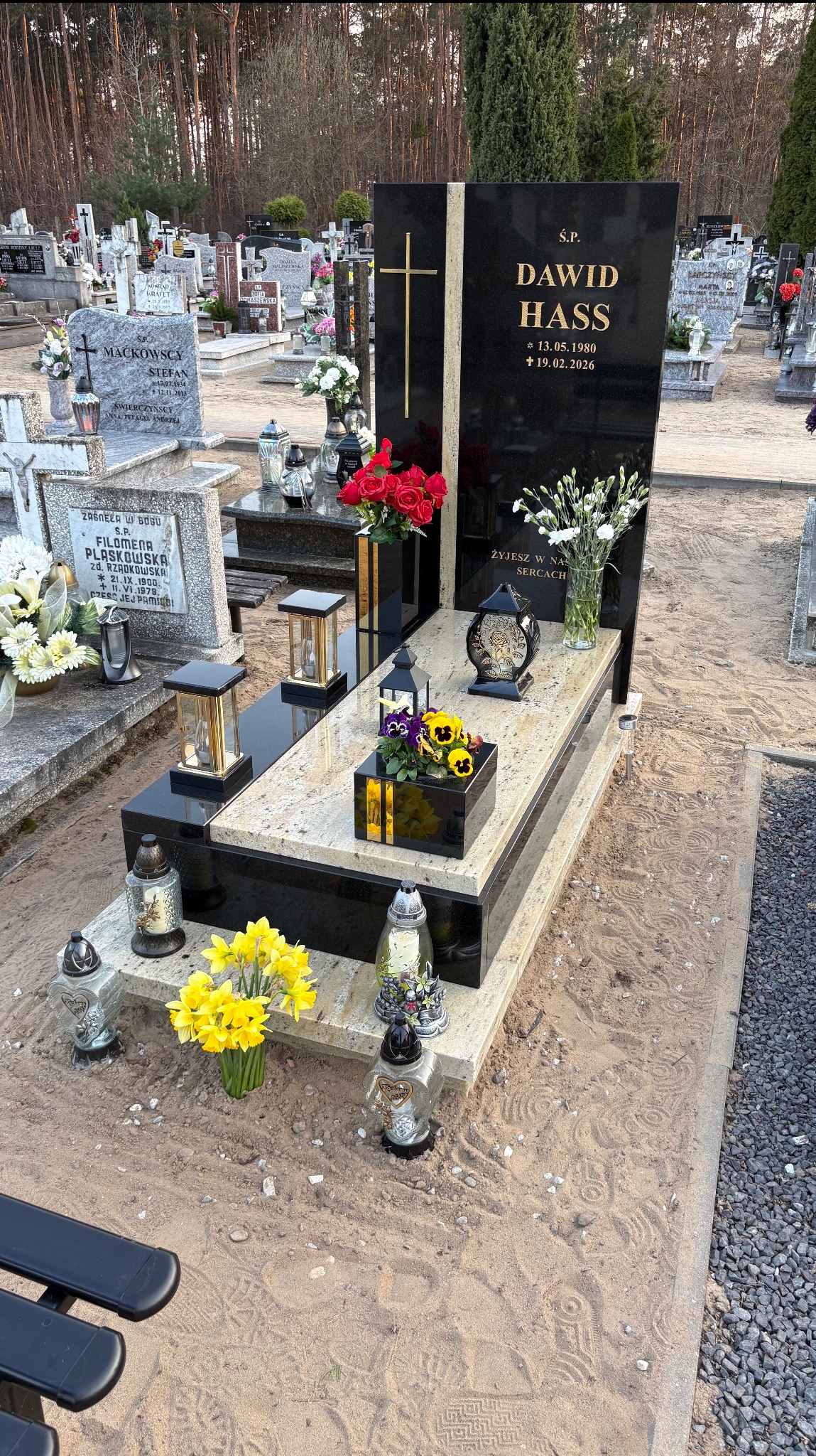 Czarny nagrobek z jasnym blatem, zdobiony złotym krzyżem i inskrypcją 'Dawid Hass', otoczony kwiatami i lampionami na cmentarzu. Widoczne ślady stóp na piasku.