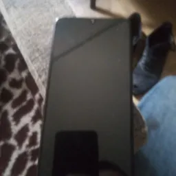 Czarny smartfon leży na kolanach osoby w dżinsach, obok widoczne buty i fragment wzorzystego dywanu. Niska jakość zdjęcia.