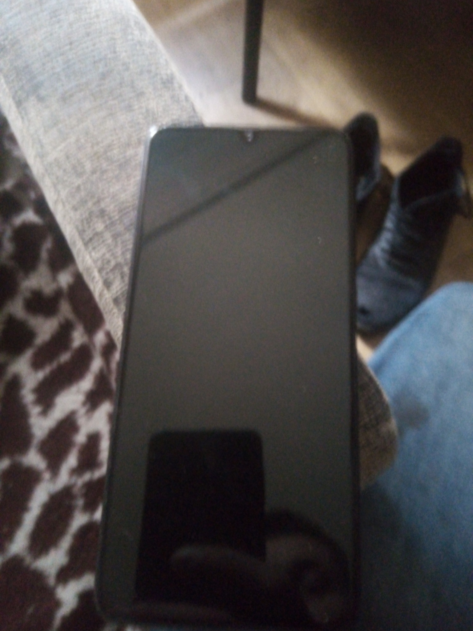 Czarny smartfon leży na kolanach osoby w dżinsach, obok widoczne buty i fragment wzorzystego dywanu. Niska jakość zdjęcia.