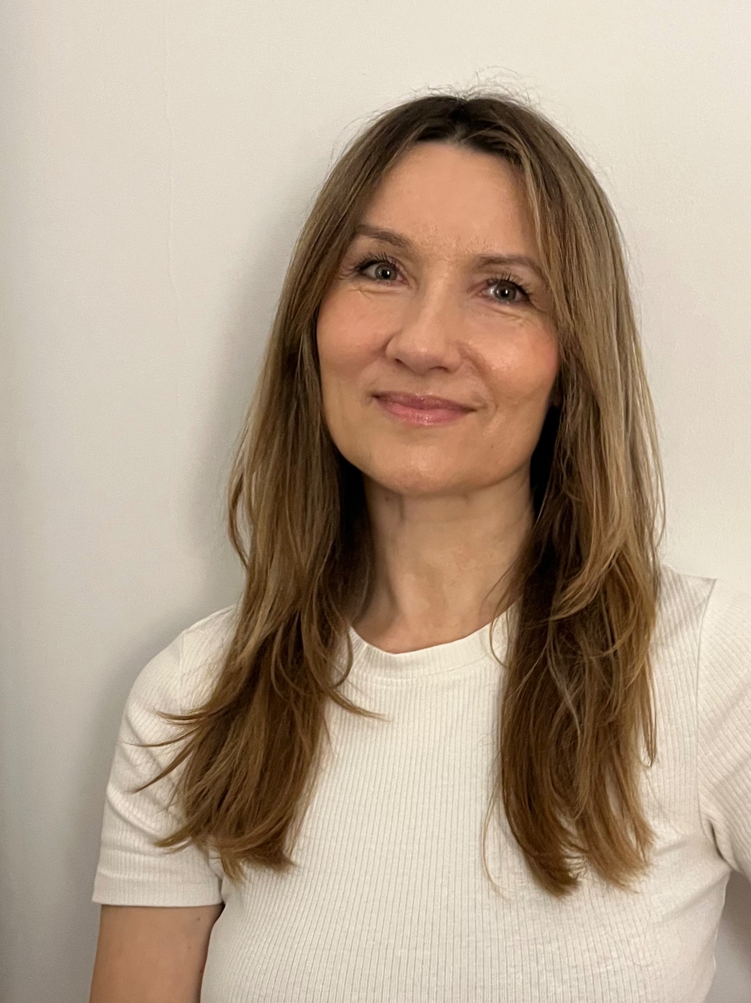 Portret uśmiechniętej kobiety o długich, brązowych włosach z delikatnym ombre, w białej bluzce na tle jasnej ściany.