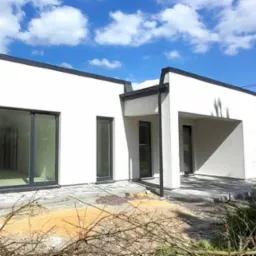 Nowoczesny, parterowy dom z białymi ścianami i czarnymi ramami okiennymi w trakcie wykańczania, z widocznym fragmentem dachu i kominem, na tle błękitnego nieba z chmurami.