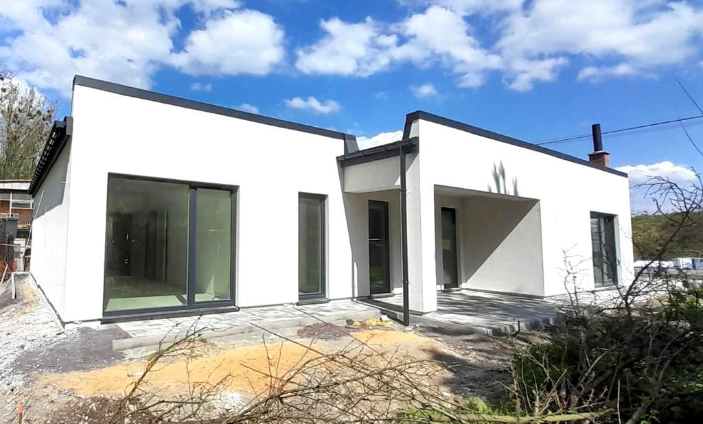 Nowoczesny, parterowy dom z białymi ścianami i czarnymi ramami okiennymi w trakcie wykańczania, z widocznym fragmentem dachu i kominem, na tle błękitnego nieba z chmurami.