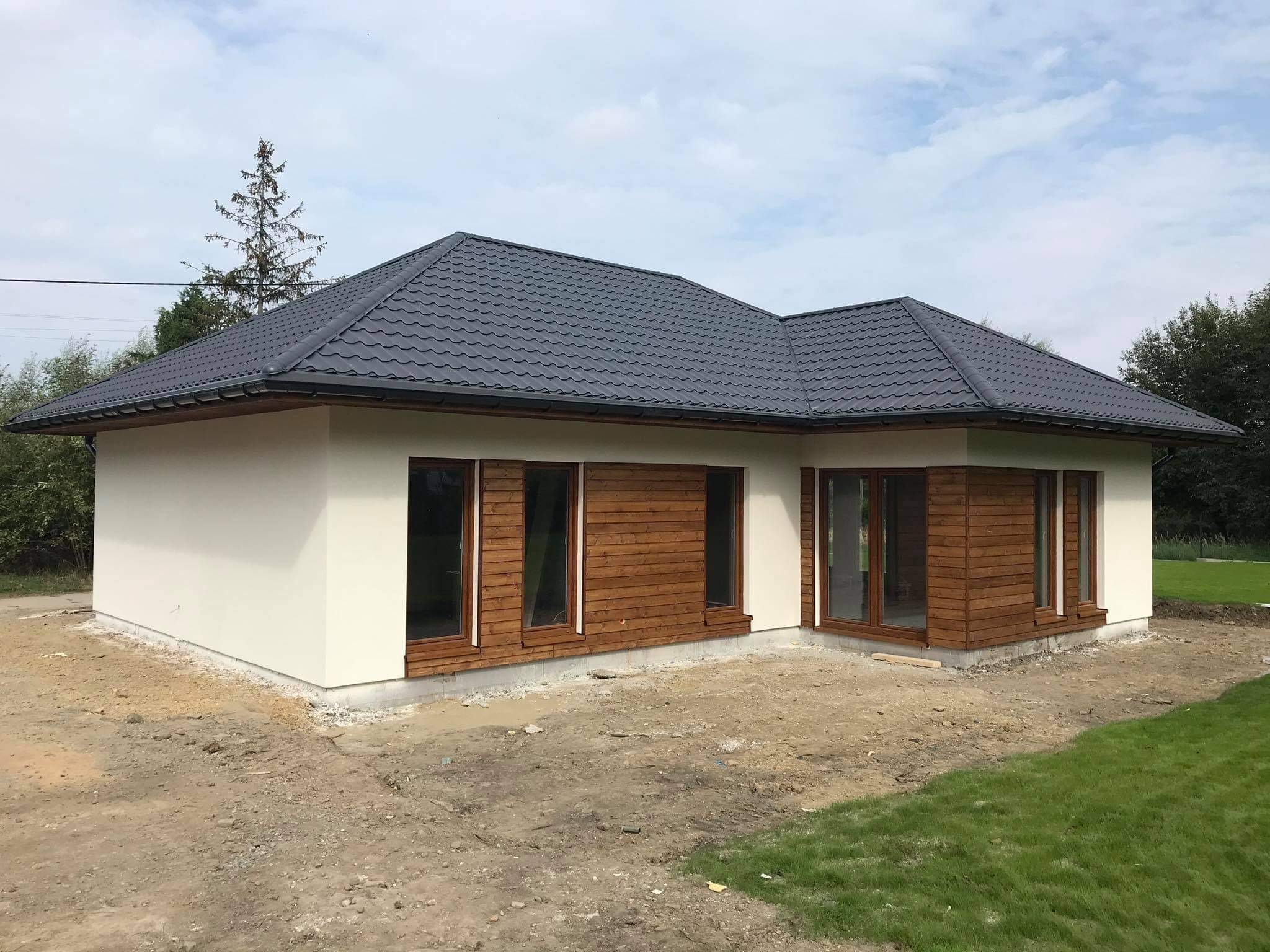 Niski parterowy dom z szarą dachówką i drewnianą elewacją w trakcie wykończenia, widoczne przygotowane podłoże pod trawnik.