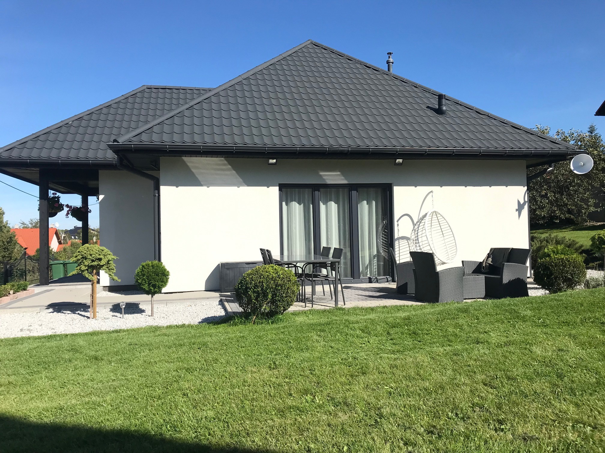 Nowoczesny, parterowy dom z szarą dachówką i białymi ścianami, z tarasem wyposażonym w meble ogrodowe i wiszące krzesło, otoczony trawnikiem i ozdobnymi krzewami w słoneczny dzień.