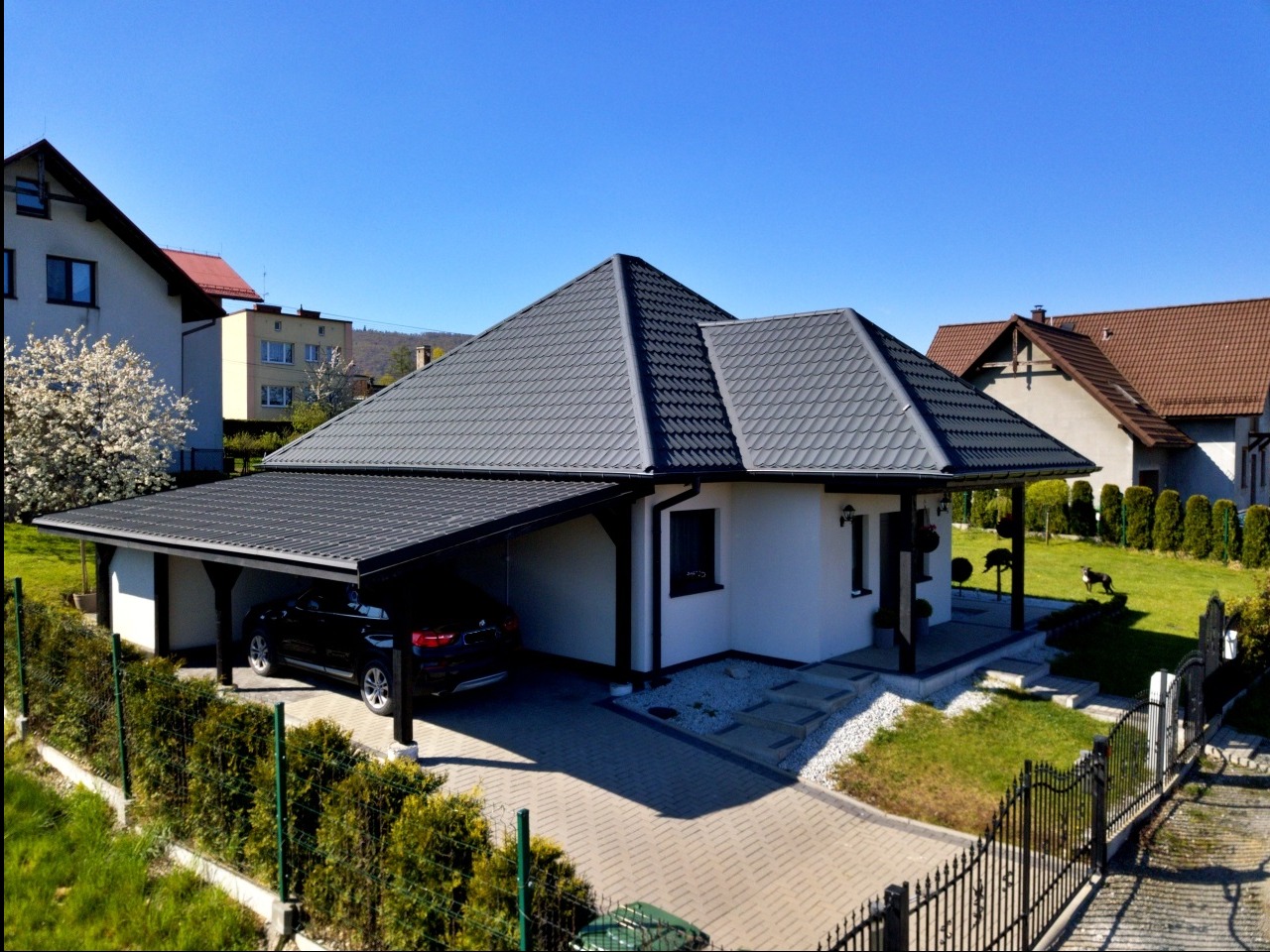 Nowoczesny dom parterowy z szarą dachówką i dobudowanym zadaszeniem na samochód, widok z góry na tle zabudowy jednorodzinnej i błękitnego nieba.