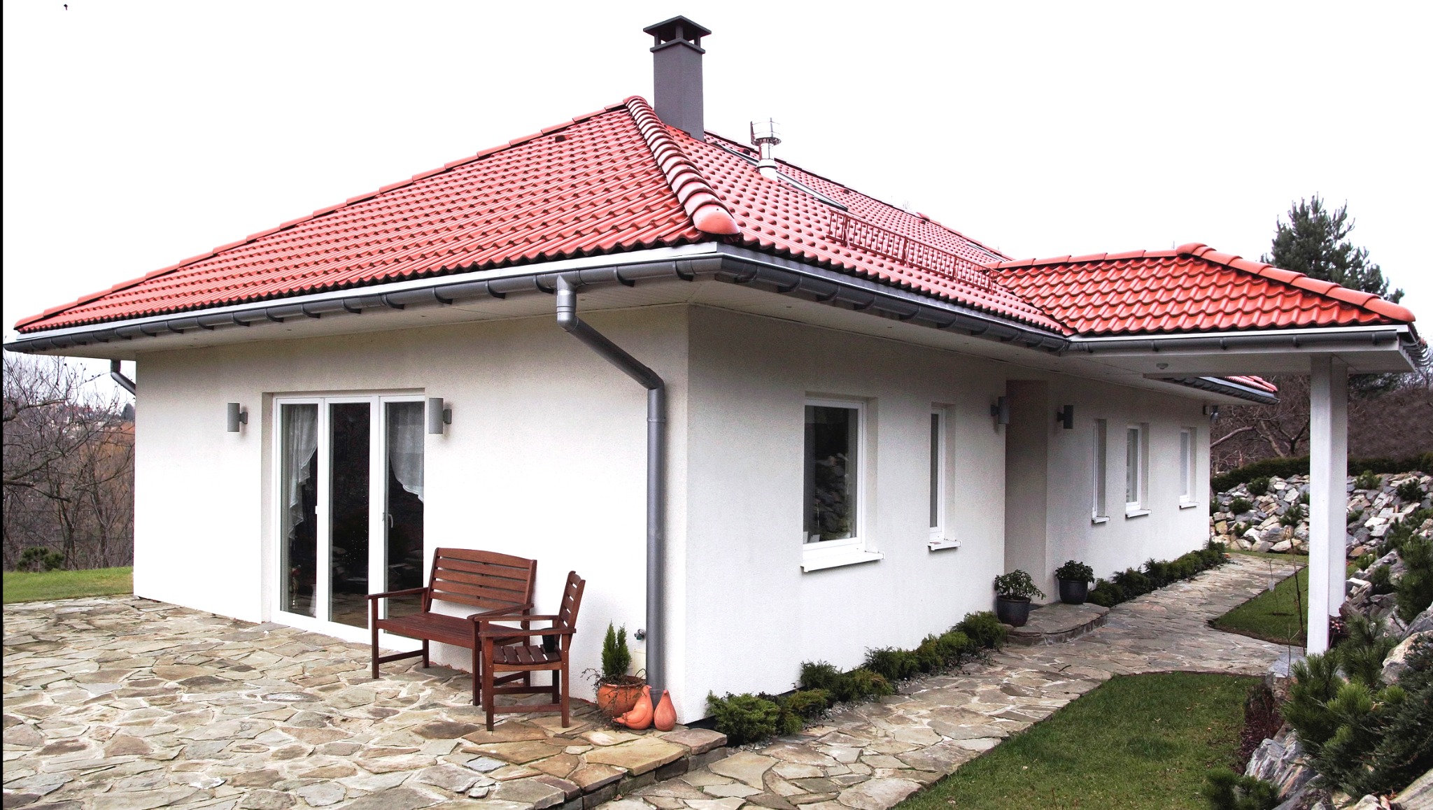 Gotowy parterowy dom z czerwoną dachówką, drewnianą ławką i kamiennym tarasem na zewnątrz.