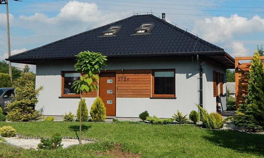 Gotowy parterowy dom z ciemną dachówką i drewnianymi elementami elewacji, otoczony trawnikiem i ozdobnymi krzewami.