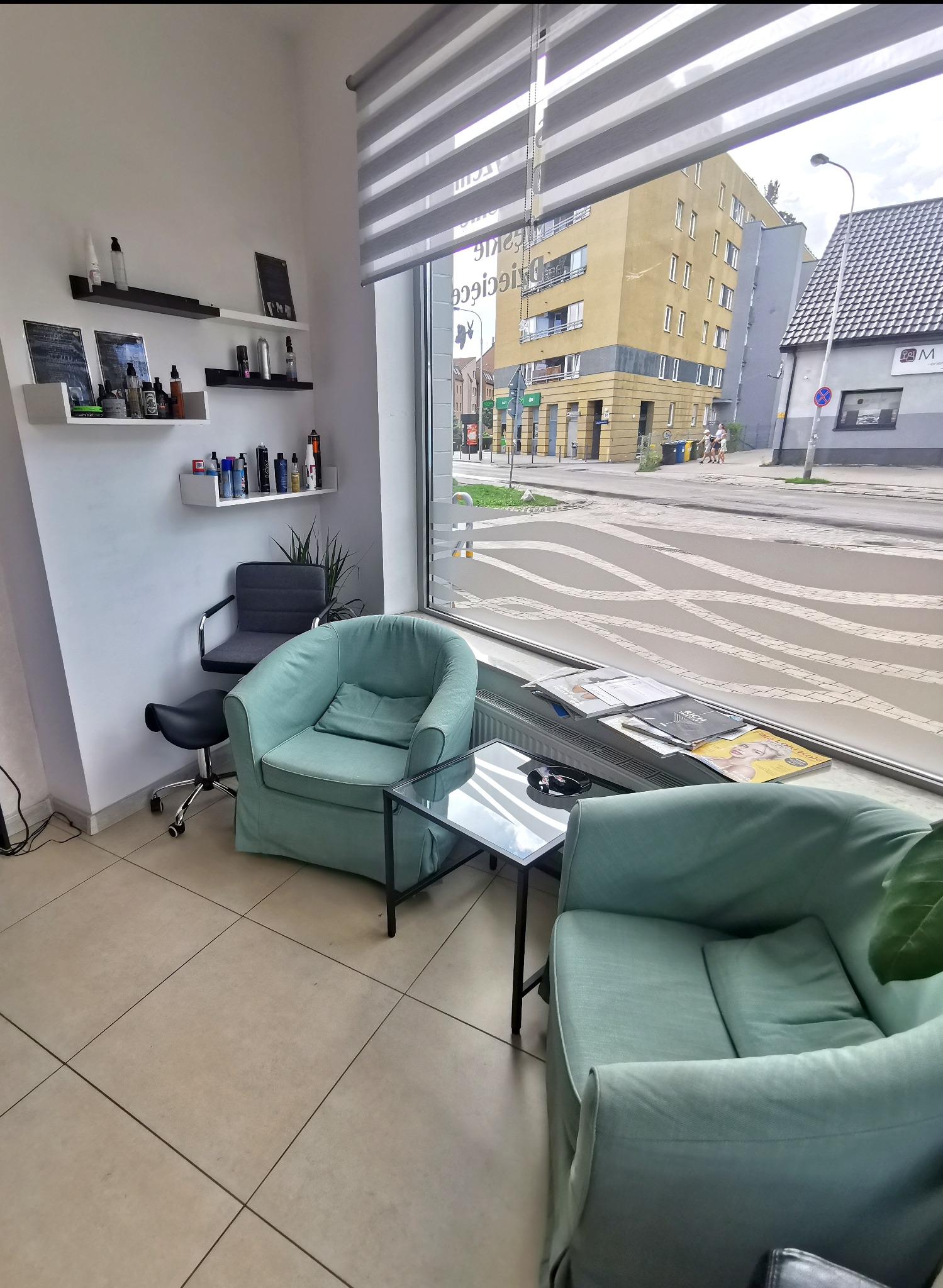 Wnętrze salonu z dwoma turkusowymi fotelami, szklanym stolikiem, półkami z kosmetykami i widokiem na ulicę przez okno z roletami.
