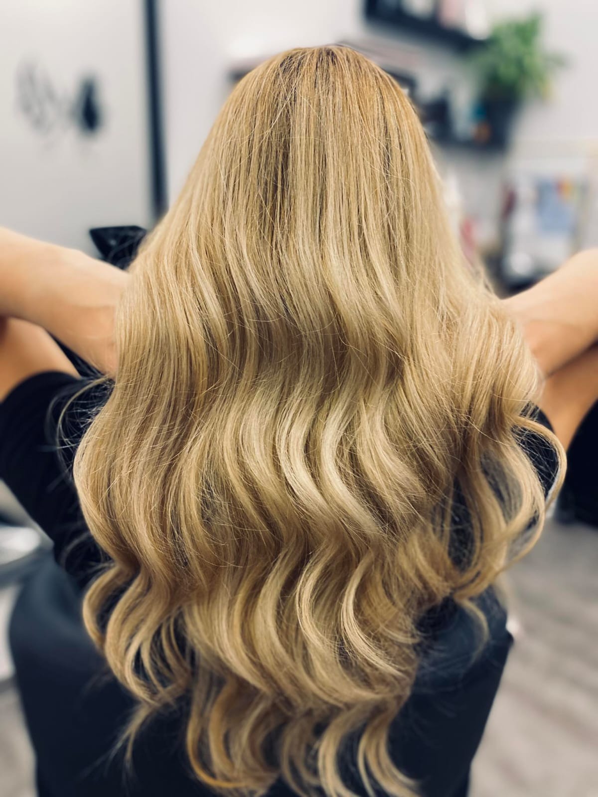 Długie, falowane, blond włosy z widocznymi refleksami, ułożone na plecach, widok z tyłu, w tle wyposażenie salonu.
