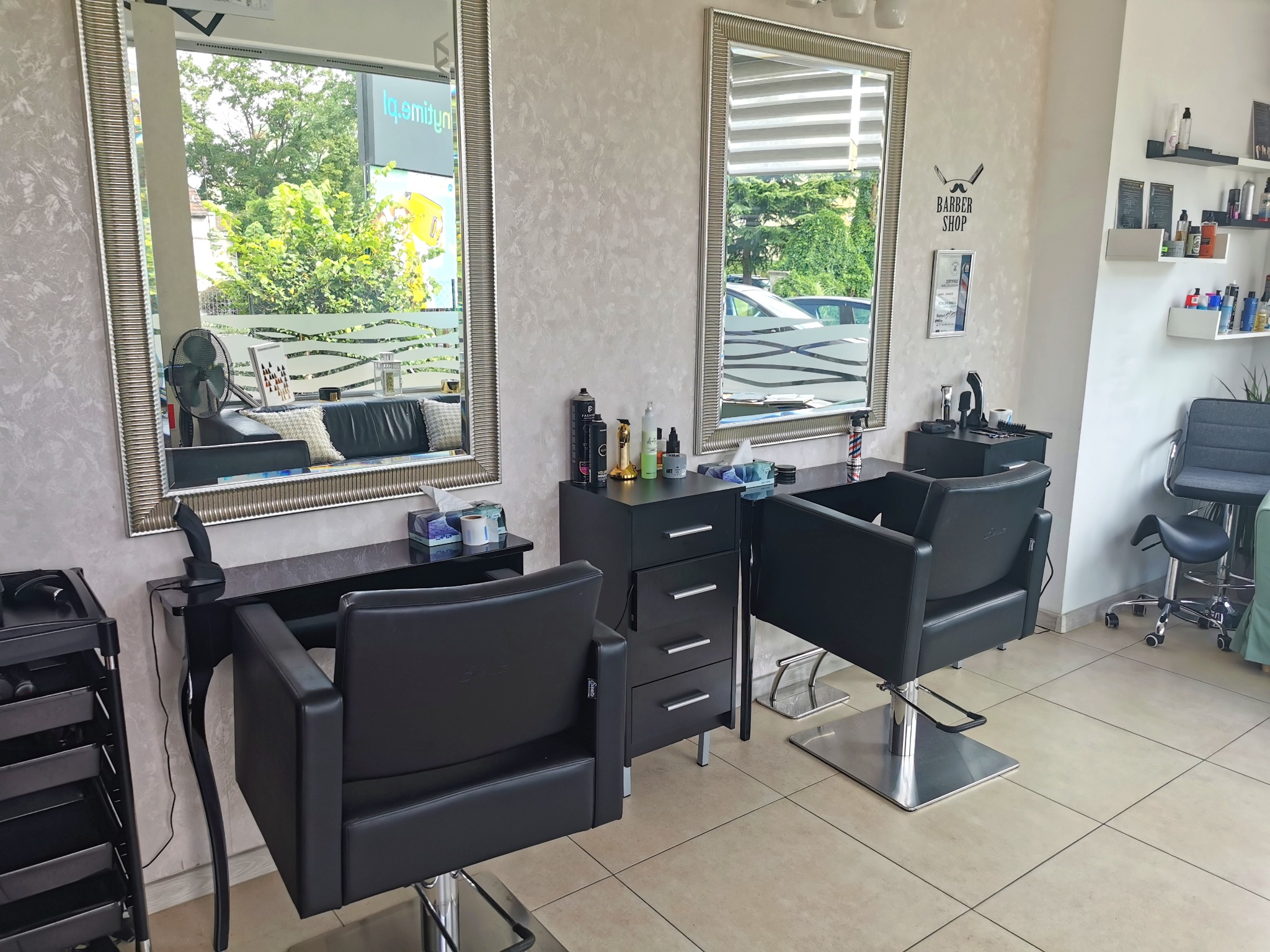 Wnętrze salonu z dwoma czarnymi fotelami fryzjerskimi, lustrami w srebrnych ramach, szafką z kosmetykami i stylizacją Barber Shop na ścianie.