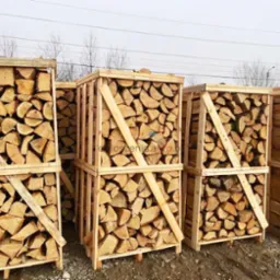 Rzęd rzędów drewnianych skrzyń wypełnionych pociętym drewnem opałowym, ustawionych na ziemi, z widocznymi śladami opon w tle i nagimi drzewami na horyzoncie.