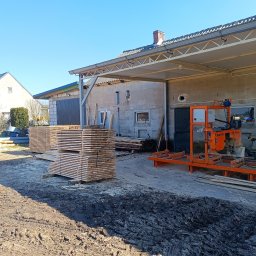 WBO Tartak - Stosy świeżo przetartej tarcicy na placu tartaku, z pomarańczową traką ramową w tle, w słoneczny dzień. Widoczne budynki gospodarcze i fragment nieba.