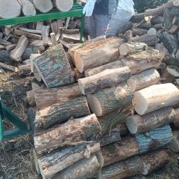 WBO Tartak - Stos pociętego drewna kominkowego, część na wózku. Pracownik w rękawicach układa polana. Widoczne słoje i korę. W tle worek z drewnem.