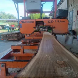 Pomarańczowa traka taśmowa Wirex CZ-1/U Mini przecina kłodę drewna. W tle stos tarcicy, budynek gospodarczy i zieleń. Widoczne trociny.