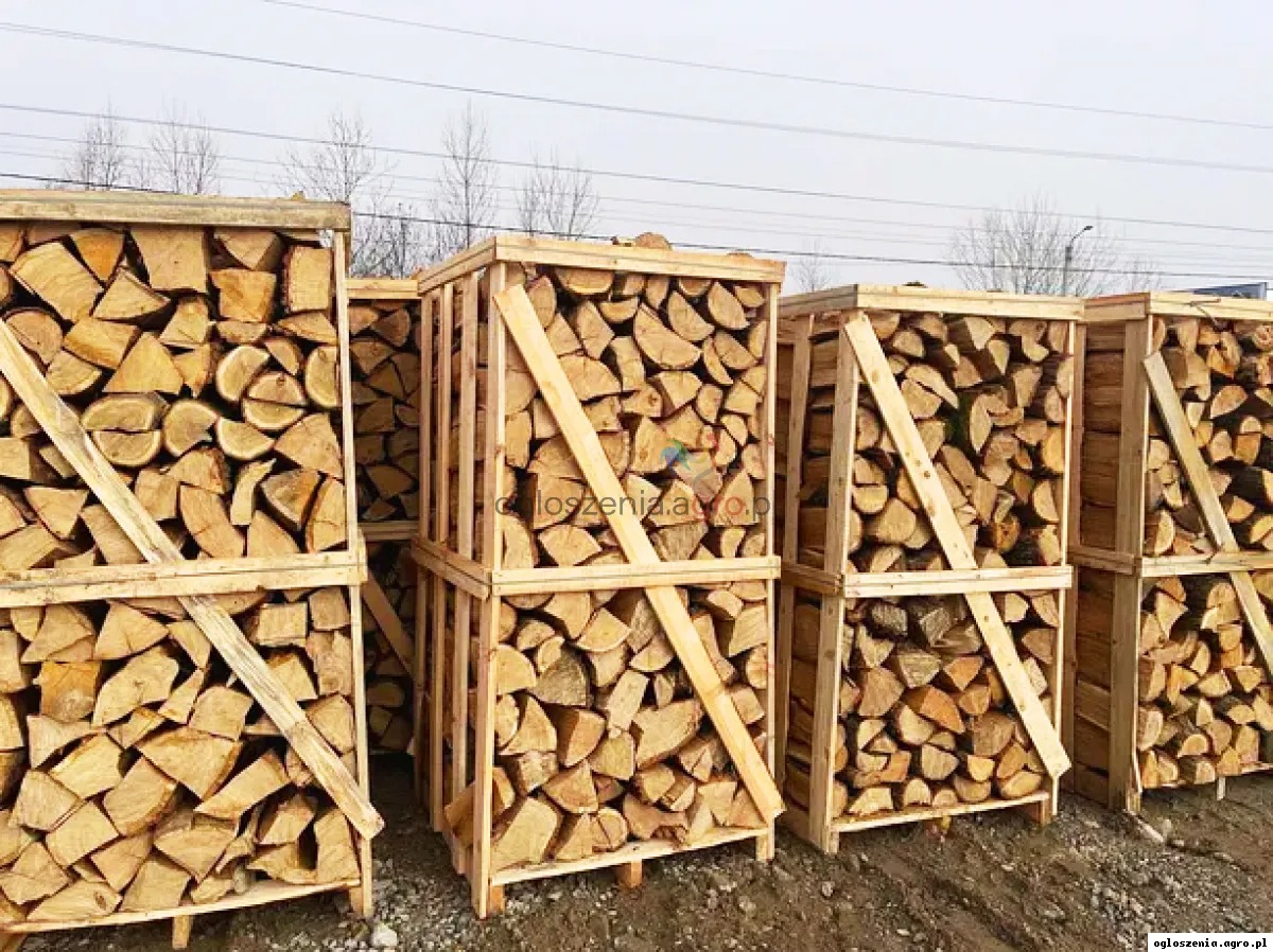 Rzęd rzędów drewnianych skrzyń wypełnionych pociętym drewnem opałowym, ustawionych na ziemi, z widocznymi śladami opon w tle i nagimi drzewami na horyzoncie.