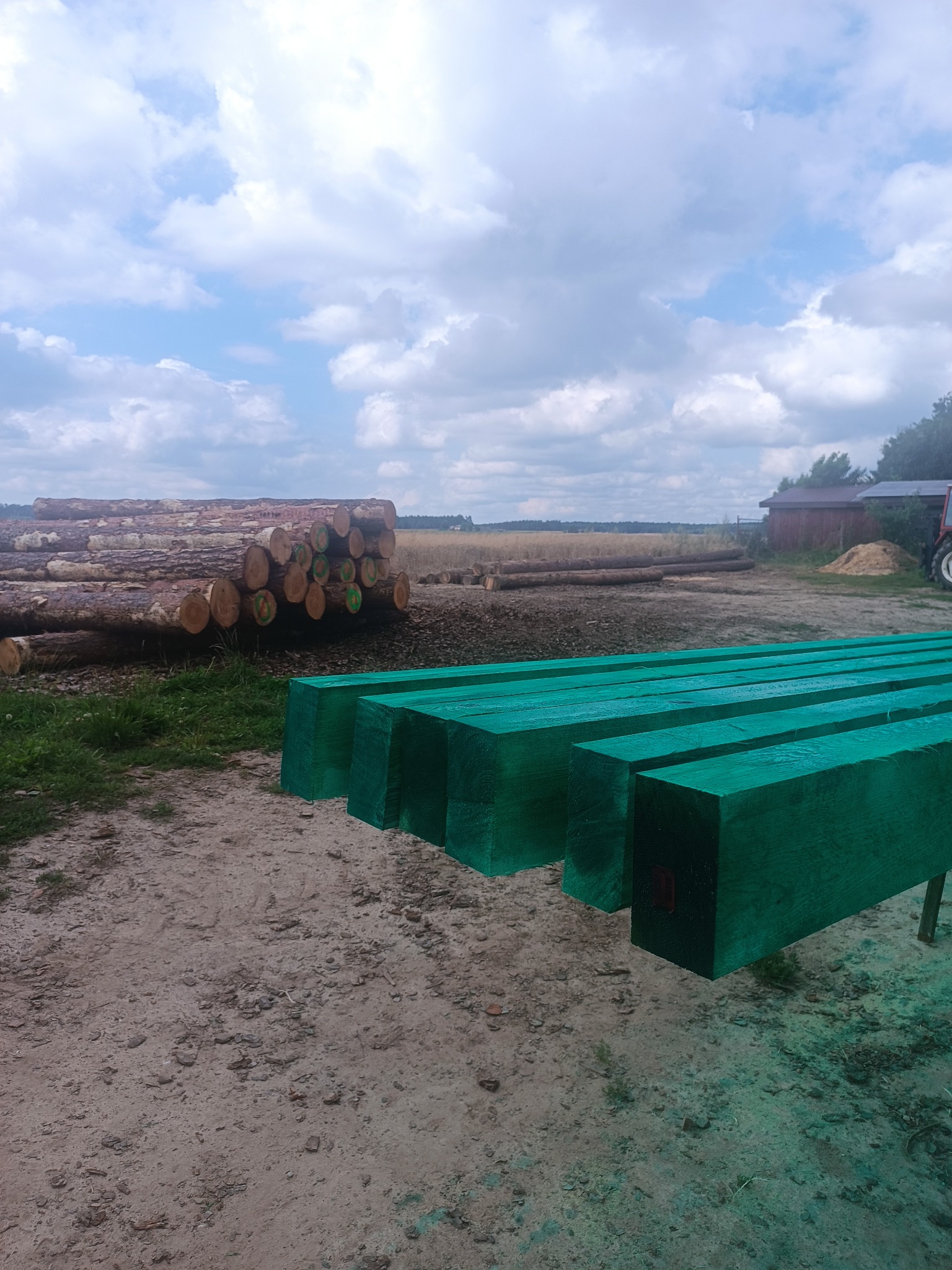 Impregnowane, zielone kantówki na tle stosu okrąglaków i krajobrazu polnego pod błękitnym niebem z chmurami. Widoczny traktor i budynek gospodarczy.