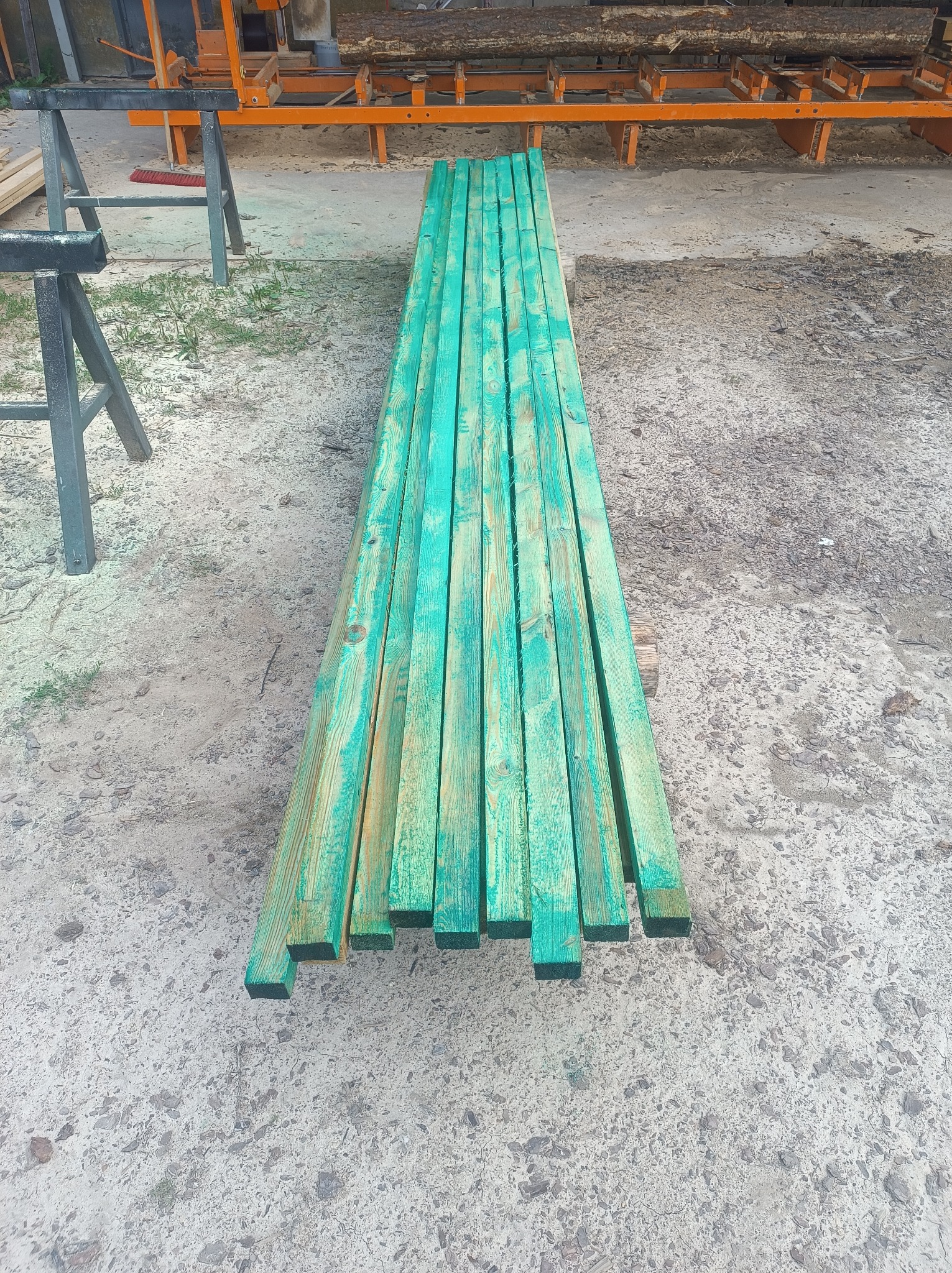 Zabezpieczone, pomalowane na zielono deski leżące na szarym, betonowym podłożu. W tle widoczny pomarańczowy element maszyny do obróbki drewna.