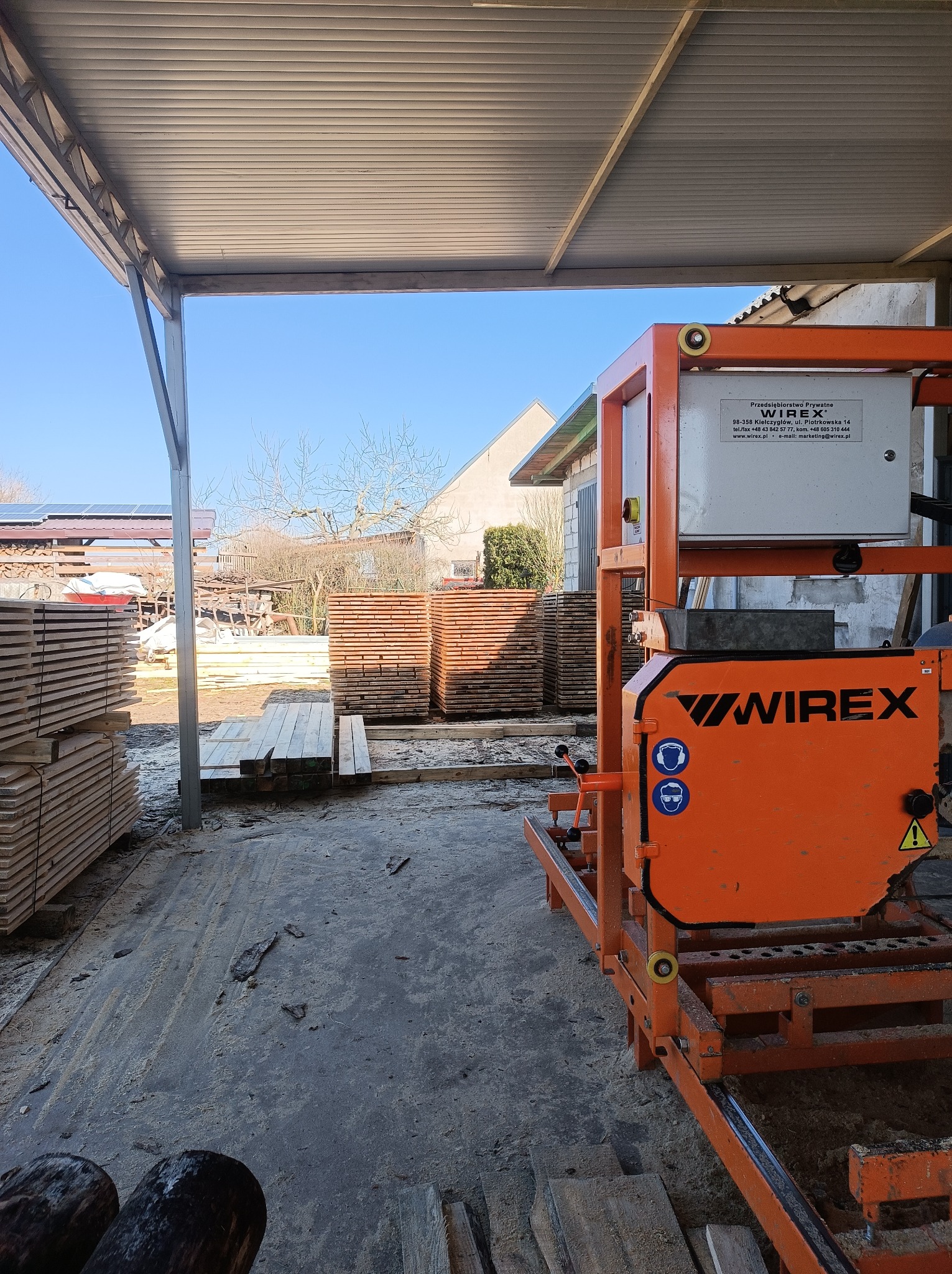 Pomarańczowa traka taśmowa Wirex w tartaku, z ułożonymi deskami w tle. Widoczne elementy konstrukcyjne budynku i fragment błękitnego nieba.