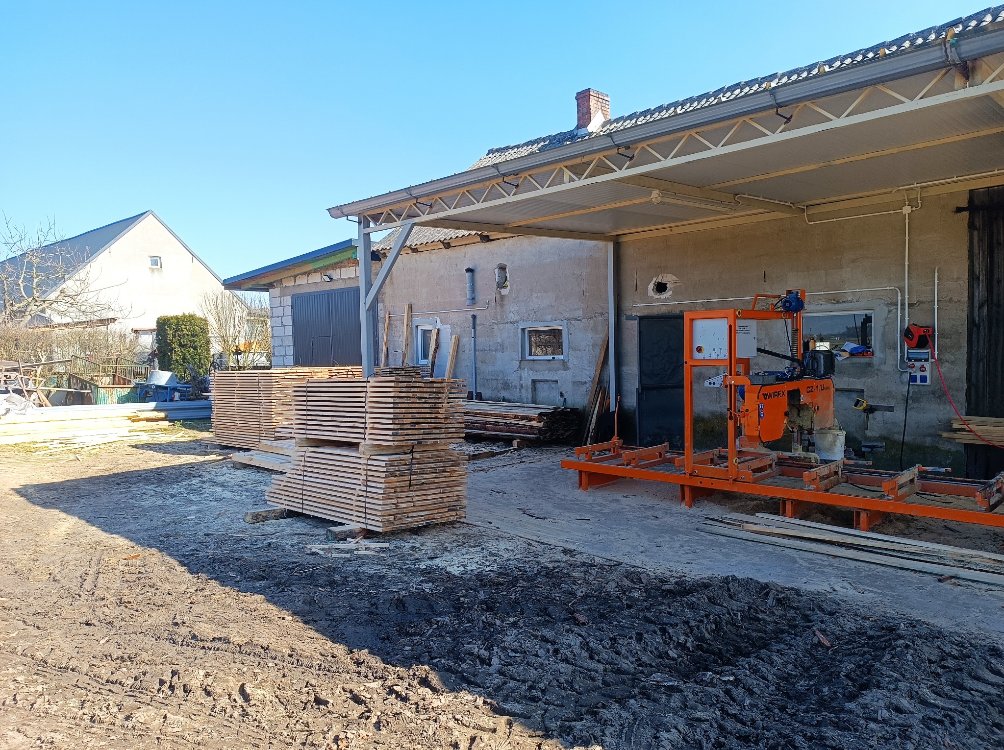 Stosy świeżo przetartej tarcicy na placu tartaku, z pomarańczową traką ramową w tle, w słoneczny dzień. Widoczne budynki gospodarcze i fragment nieba.