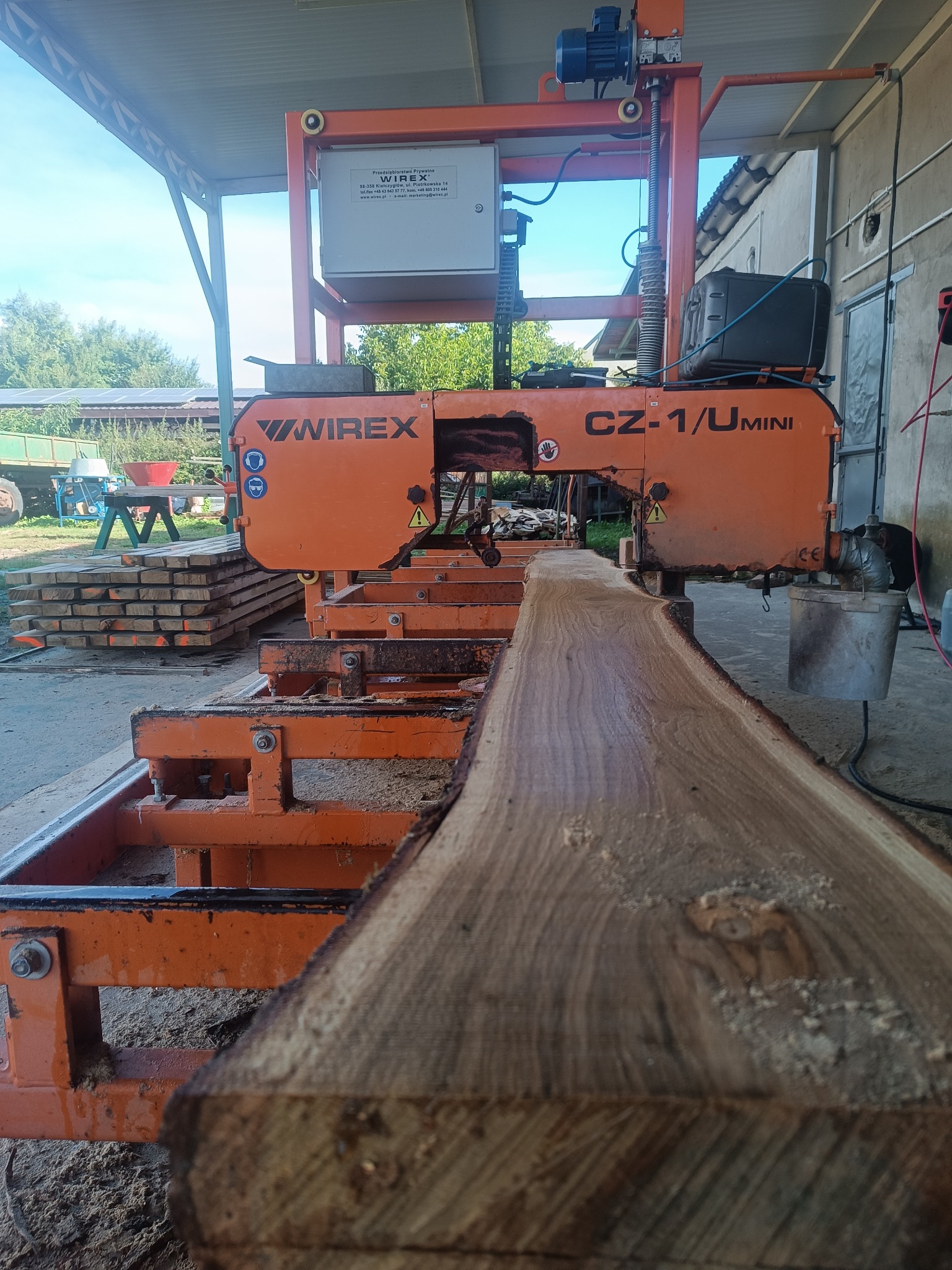 Pomarańczowa traka taśmowa Wirex CZ-1/U Mini przecina kłodę drewna. W tle stos tarcicy, budynek gospodarczy i zieleń. Widoczne trociny.