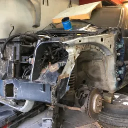 Demontaż i naprawa karoserii samochodu: widoczny fragment nadkola z licznymi otworami po spawaniu, odsłonięty układ zawieszenia i tarcza hamulcowa, puszka z farbą na masce, auto podniesione...