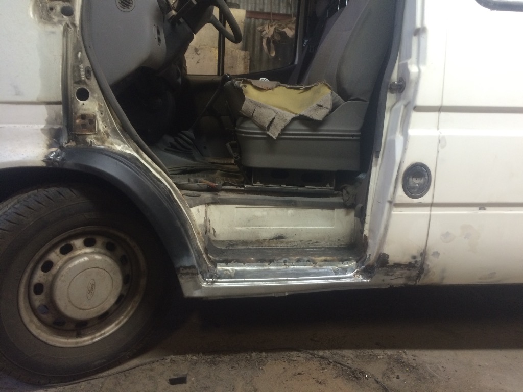 Biały samochód dostawczy z widocznymi śladami napraw blacharskich i spawalniczych w okolicy progu i nadkola, z otwartymi drzwiami kierowcy.