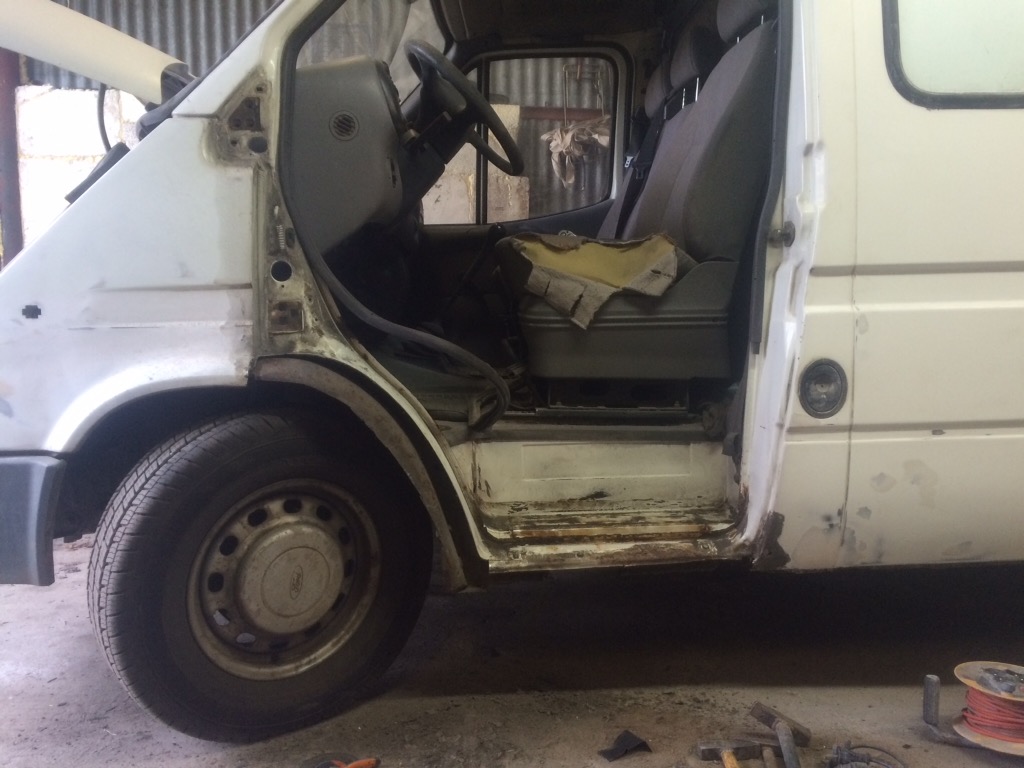 Biały van w trakcie naprawy blacharskiej: widoczne uszkodzenia karoserii, otwarte drzwi, narzędzia na podłodze.