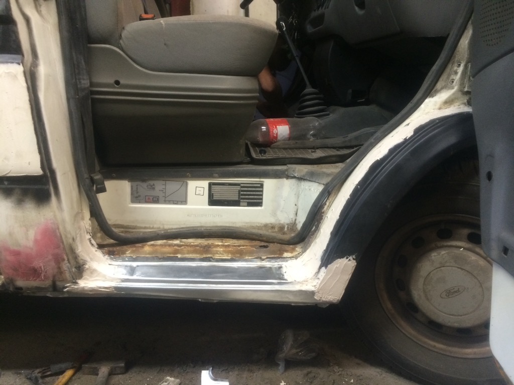 Naprawa progu i nadkola białego samochodu dostawczego, widoczne ślady korozji i szpachli, wnętrze kabiny z butelką wody